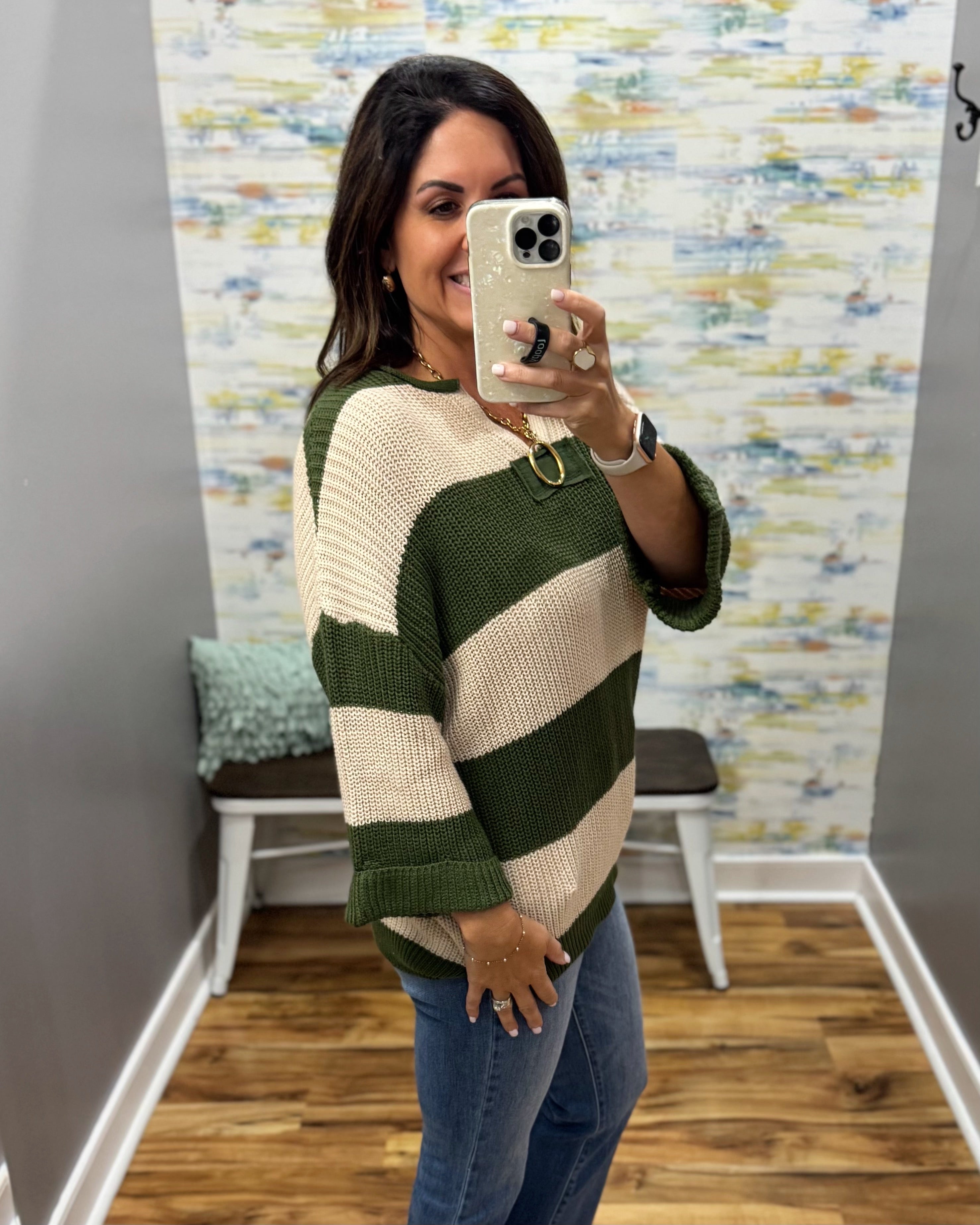 A woman takes a mirror selfie wearing a chunky knit sweater with wide horizontal stripes in dark green and cream, paired with blue jeans.