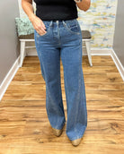 A person wearing mica wide-leg jeans, a black top, and tan pointed-toe heels, standing on a wood floor in an indoor room with a bench and abstract wall art in the background.
