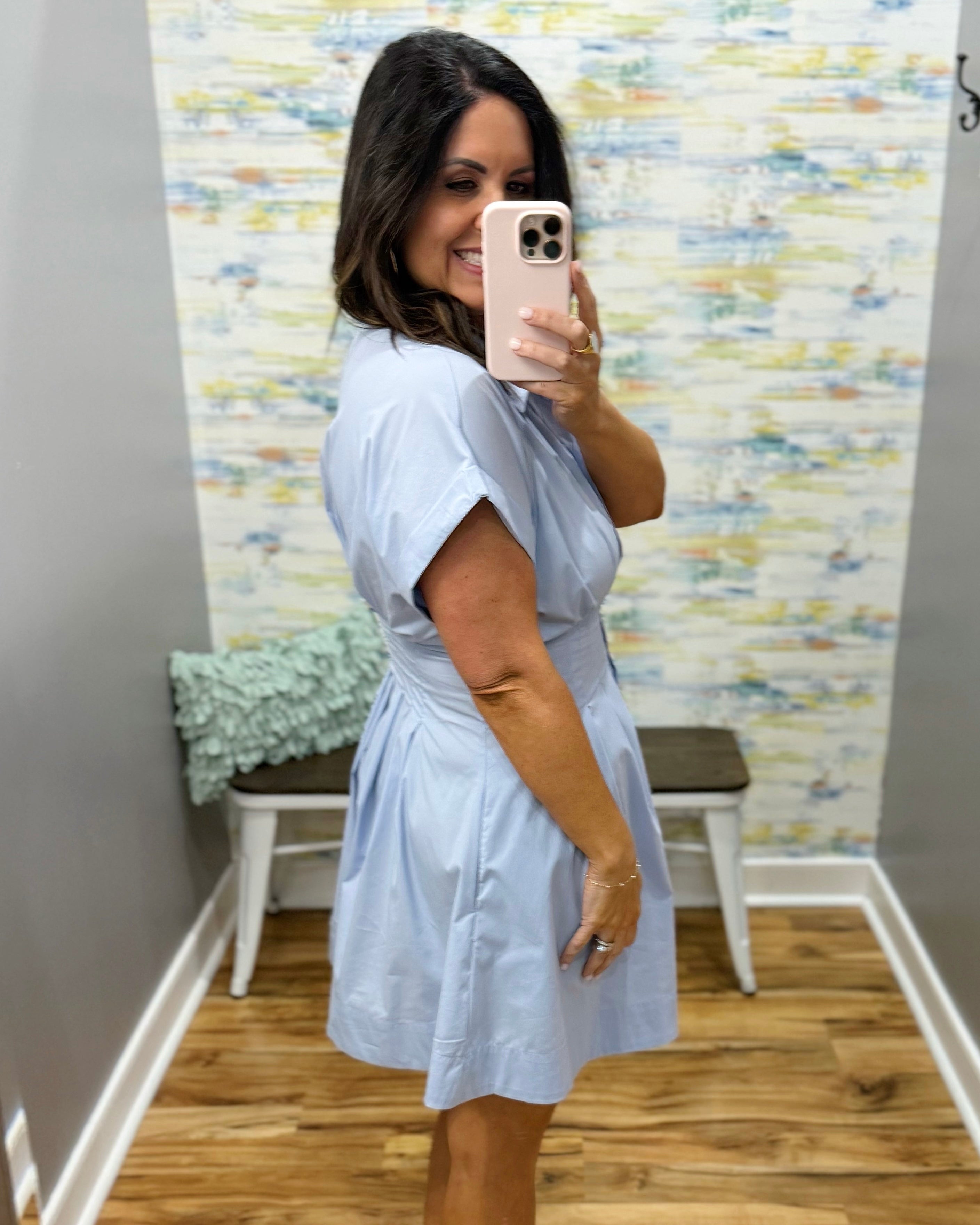 Woman taking a mirror selfie wearing a light blue dress in a room with a textured wall.