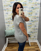 A woman takes a mirror selfie wearing a light gray short-sleeve knit sweater vest and dark blue jeans in a room with patterned wallpaper and a bench.