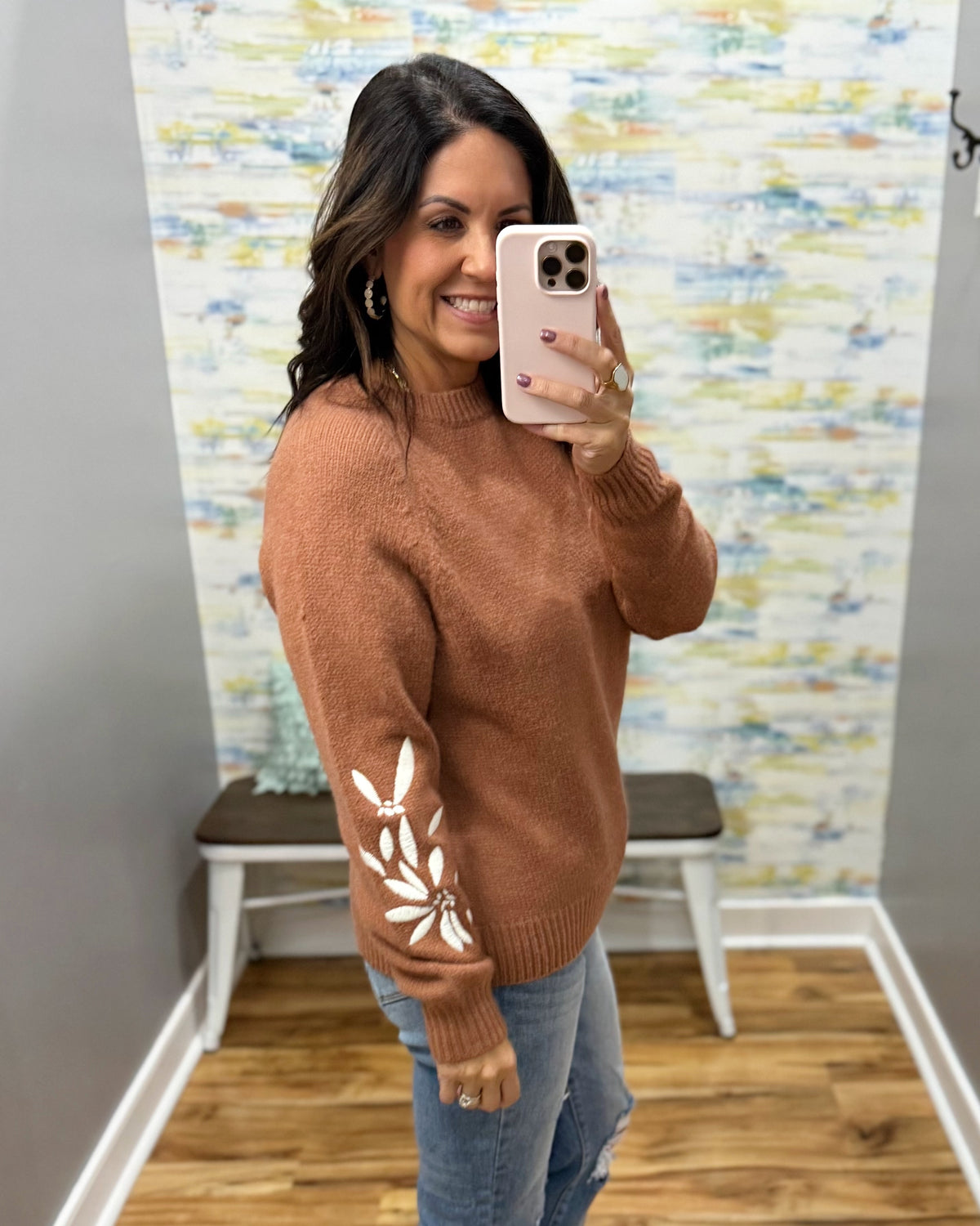 A woman takes a mirror selfie wearing a rust-brown crewneck sweater with white floral embroidery on the sleeve, paired with light-wash blue jeans.