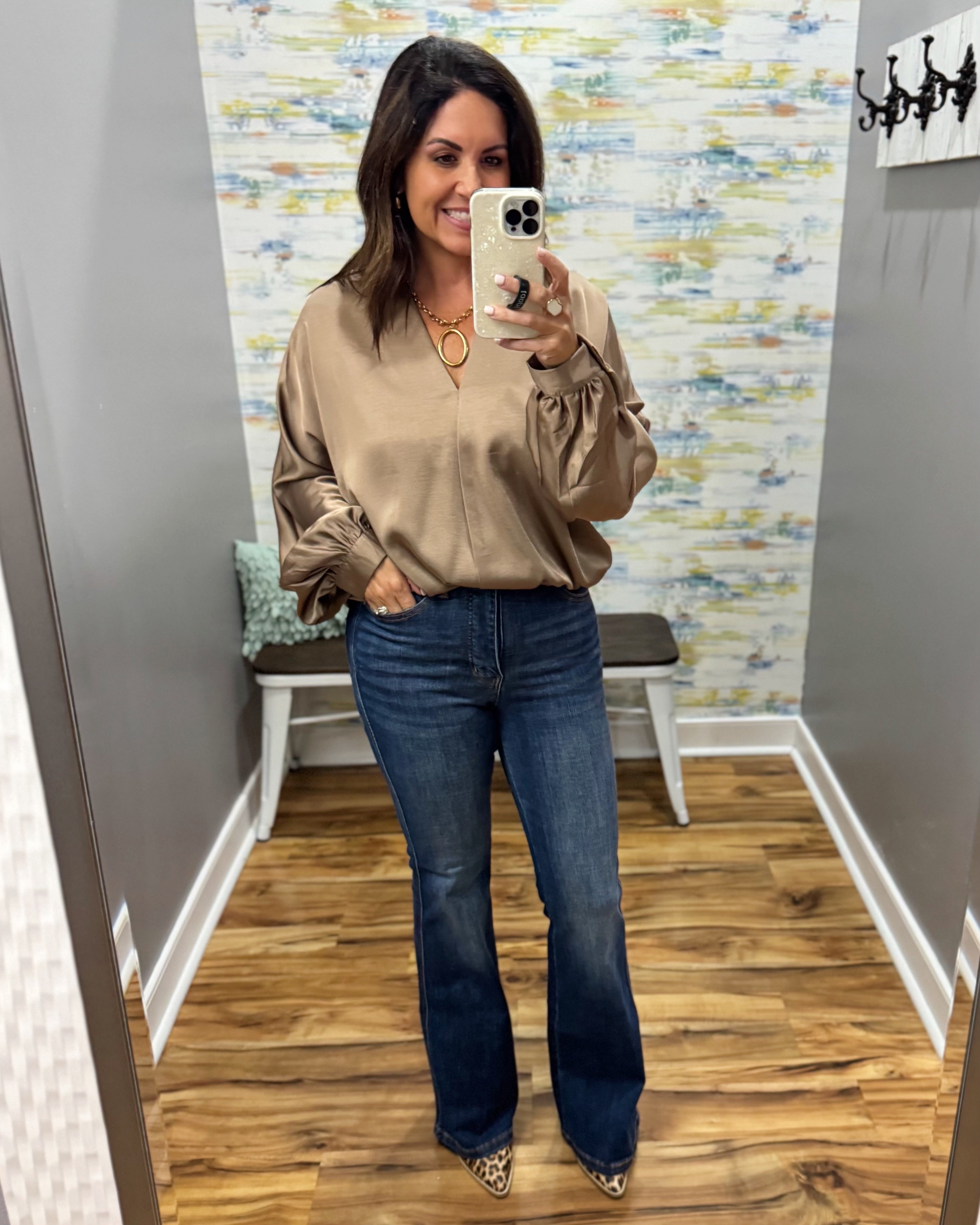 A woman takes a mirror selfie in a fitting room wearing a beige long-sleeve blouse with gathered sleeves and dark blue flare jeans.