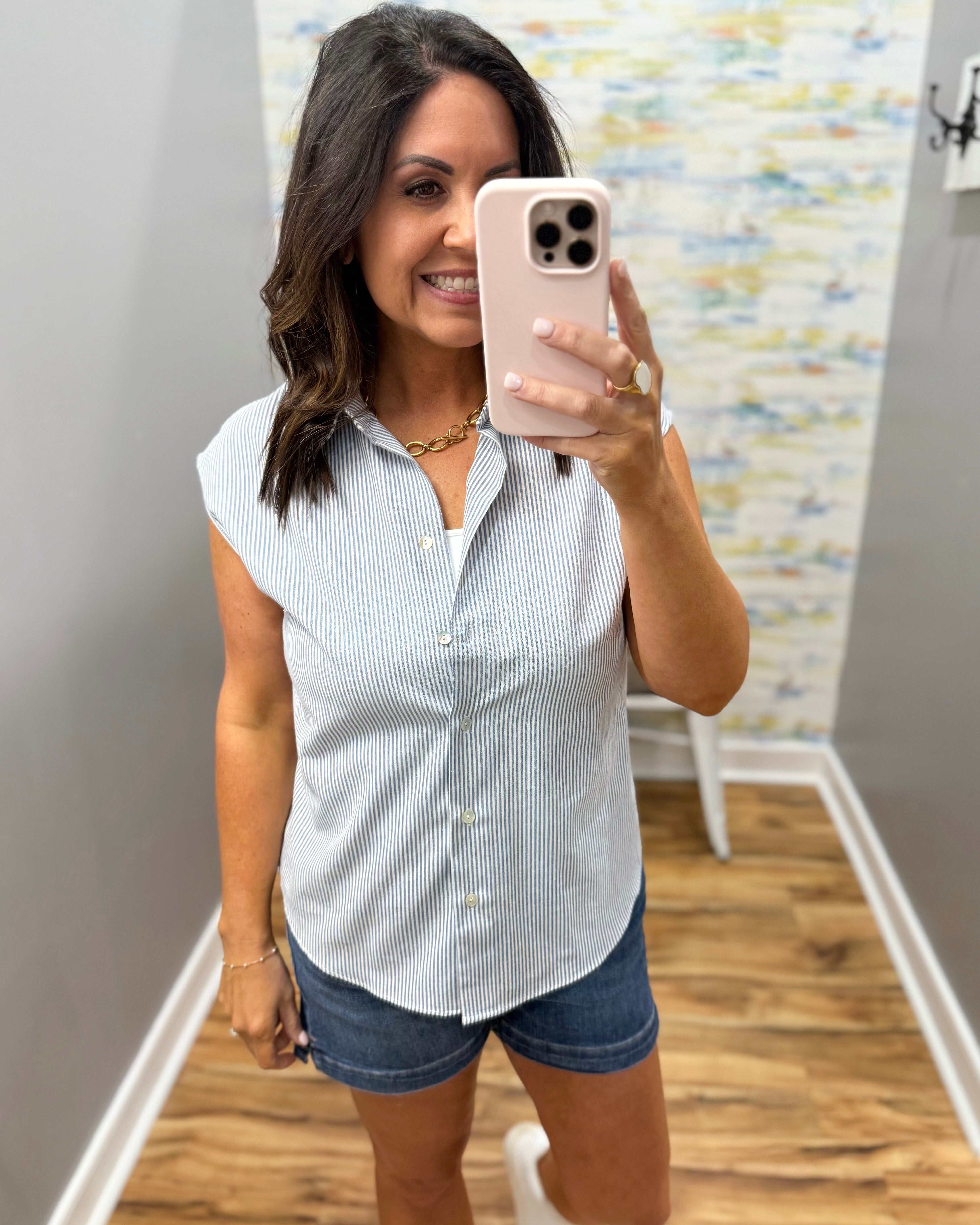 Woman taking a mirror selfie wearing a white button-up shirt and denim shorts in a room with a colorful wall.