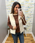 A woman takes a mirror selfie wearing a cream-colored sleeveless puffer vest over a brown long-sleeve top and blue jeans.