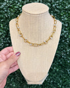 Chansuttpearls Jennifer gold chain necklace with small clear crystal accents, displayed on a beige fabric jewelry bust. A hand with purple nail polish is visible on the left side, holding the bust against a green leaf hedge background.