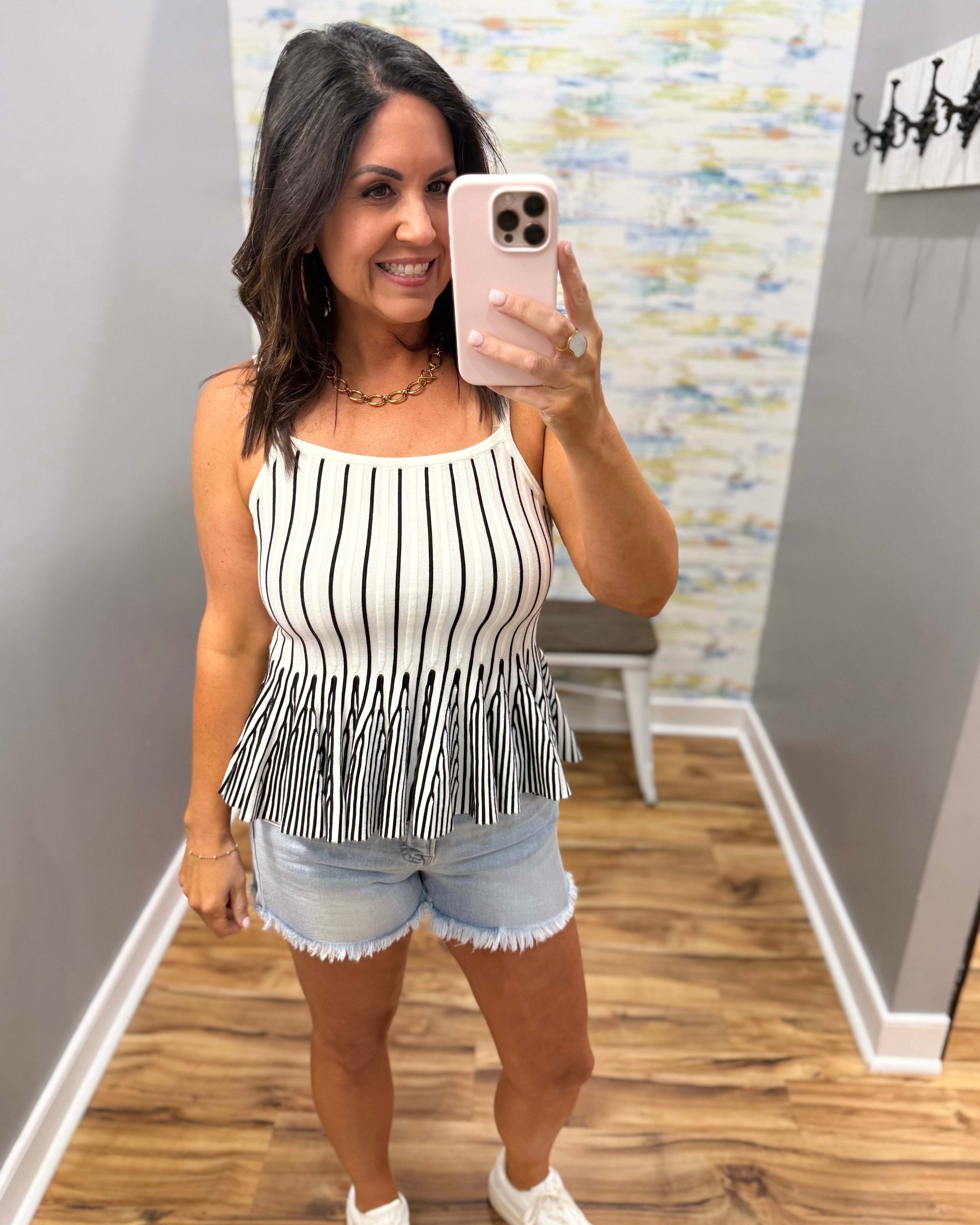 Woman taking a mirror selfie wearing a black and white striped top with fringe and denim shorts.