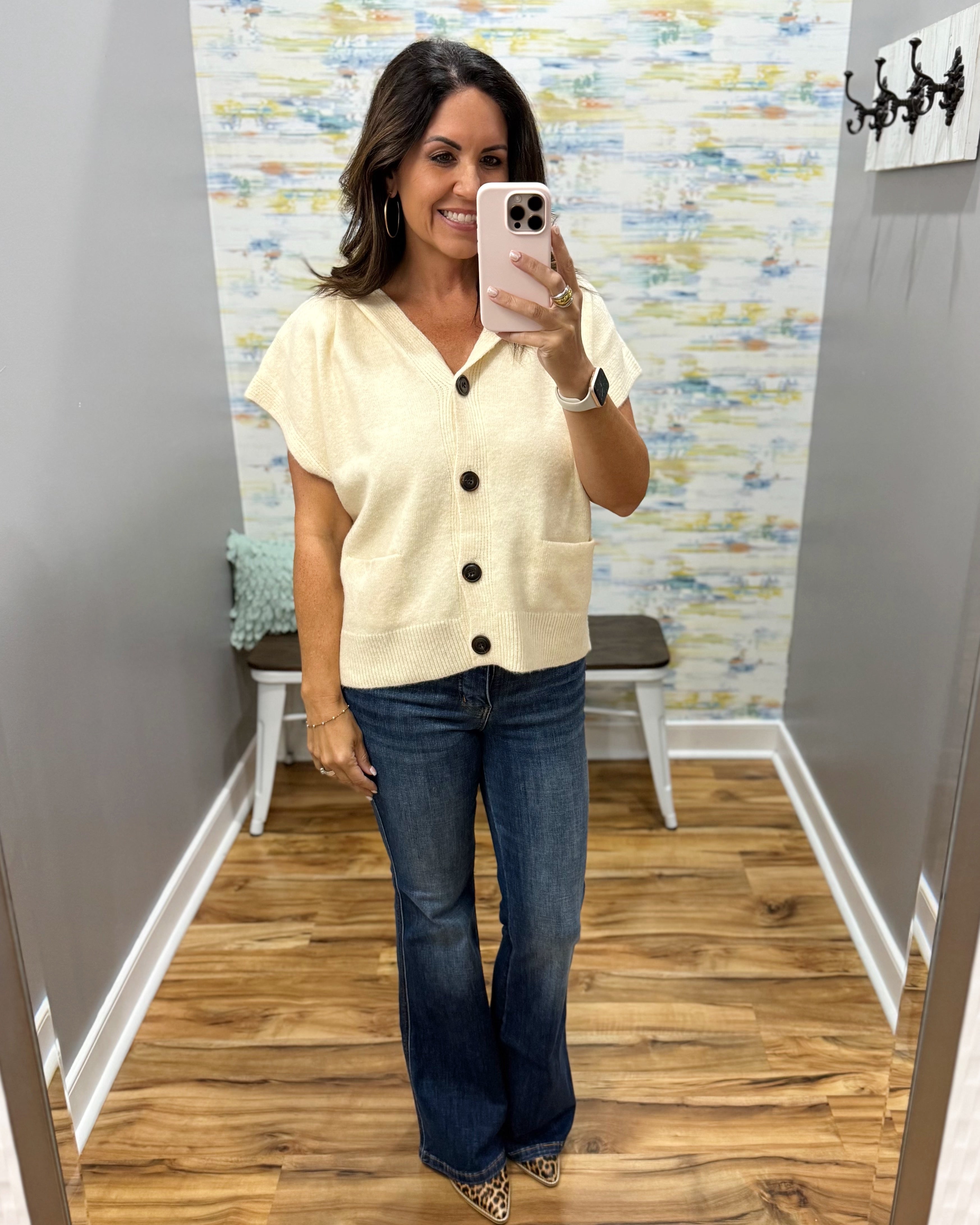 A woman takes a mirror selfie wearing a cream short-sleeve button-front cardigan and blue flare jeans in a fitting room with wood floors and a patterned wallpaper background.