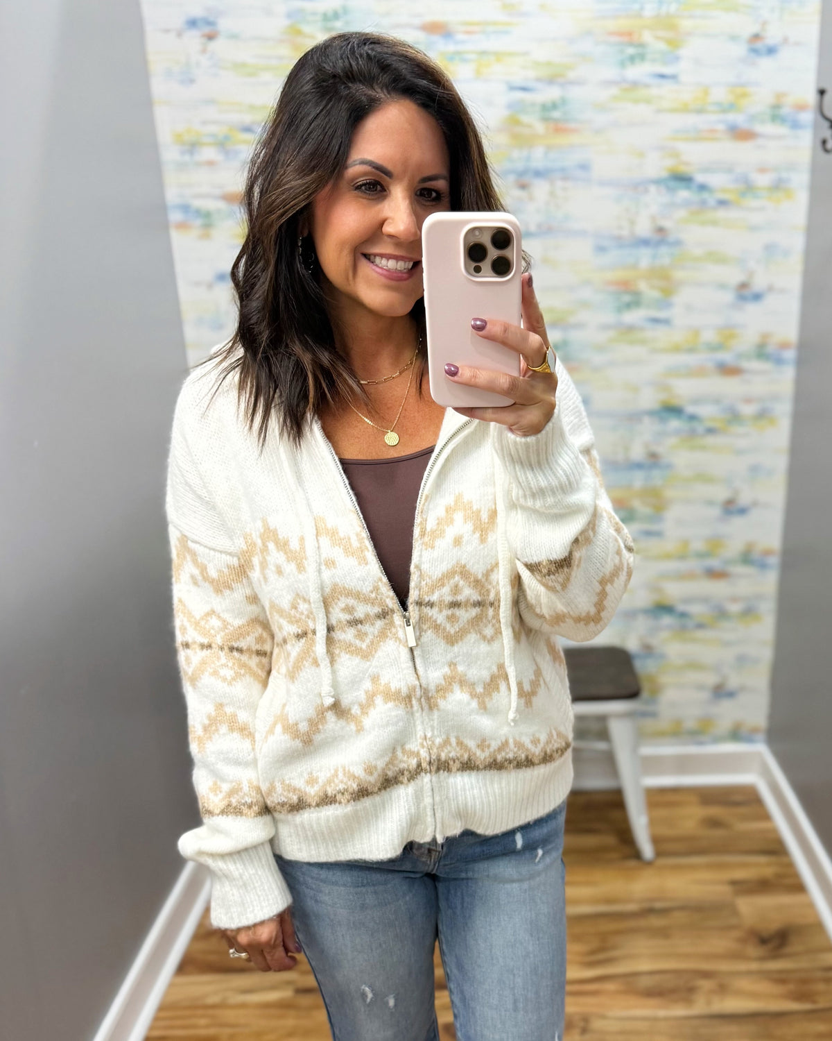 A woman taking a mirror selfie wearing a cream and tan Fair Isle knit zip-up cardigan over a brown top and blue jeans.