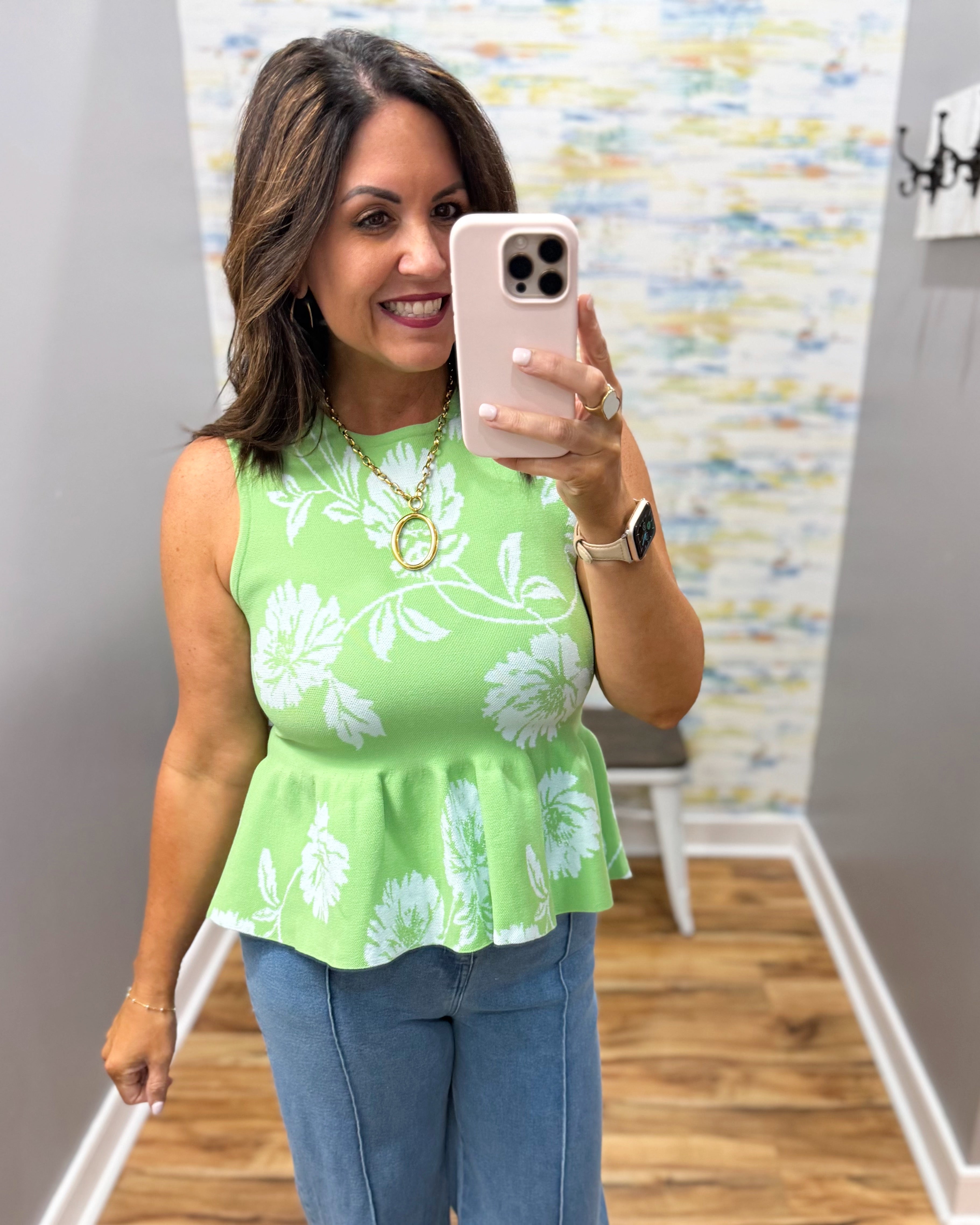 Woman taking a mirror selfie wearing a green floral peplum top and blue jeans.