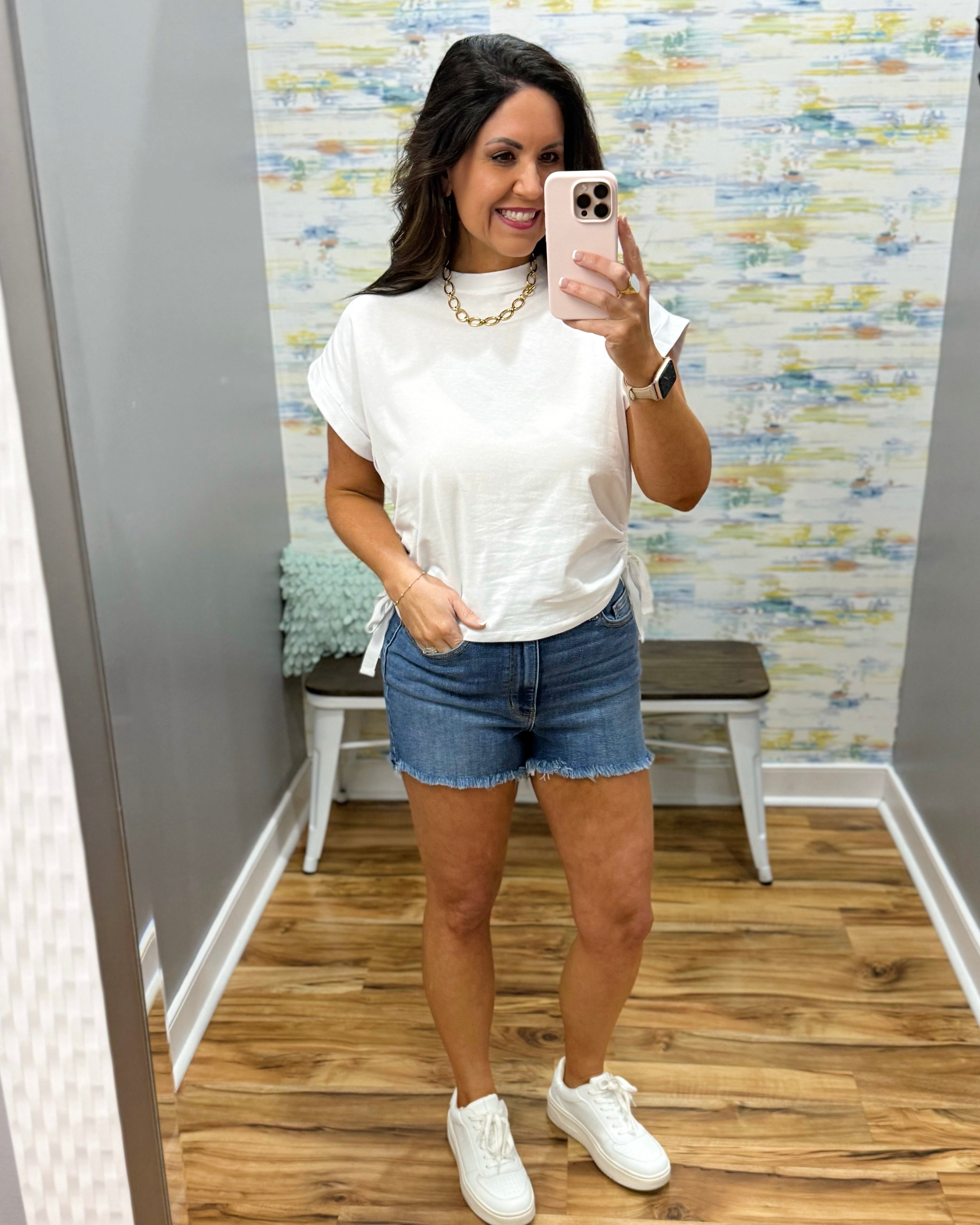 Woman taking a mirror selfie wearing a white t-shirt, denim shorts, and white sneakers in a room with a patterned wall.