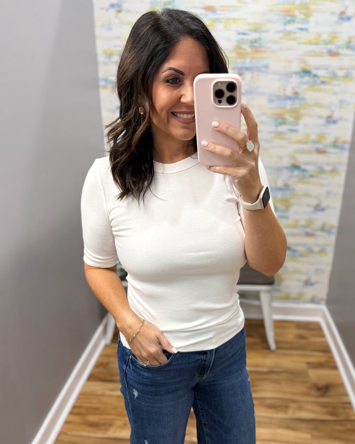 A woman taking a mirror selfie wearing a fitted white short-sleeve crewneck top and blue jeans in an indoor room with wood flooring and a patterned wall.