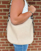 A woman wearing a white tank top and blue jeans carries a large beige suede shoulder bag with black and silver chain strap details, set against a red brick wall background.