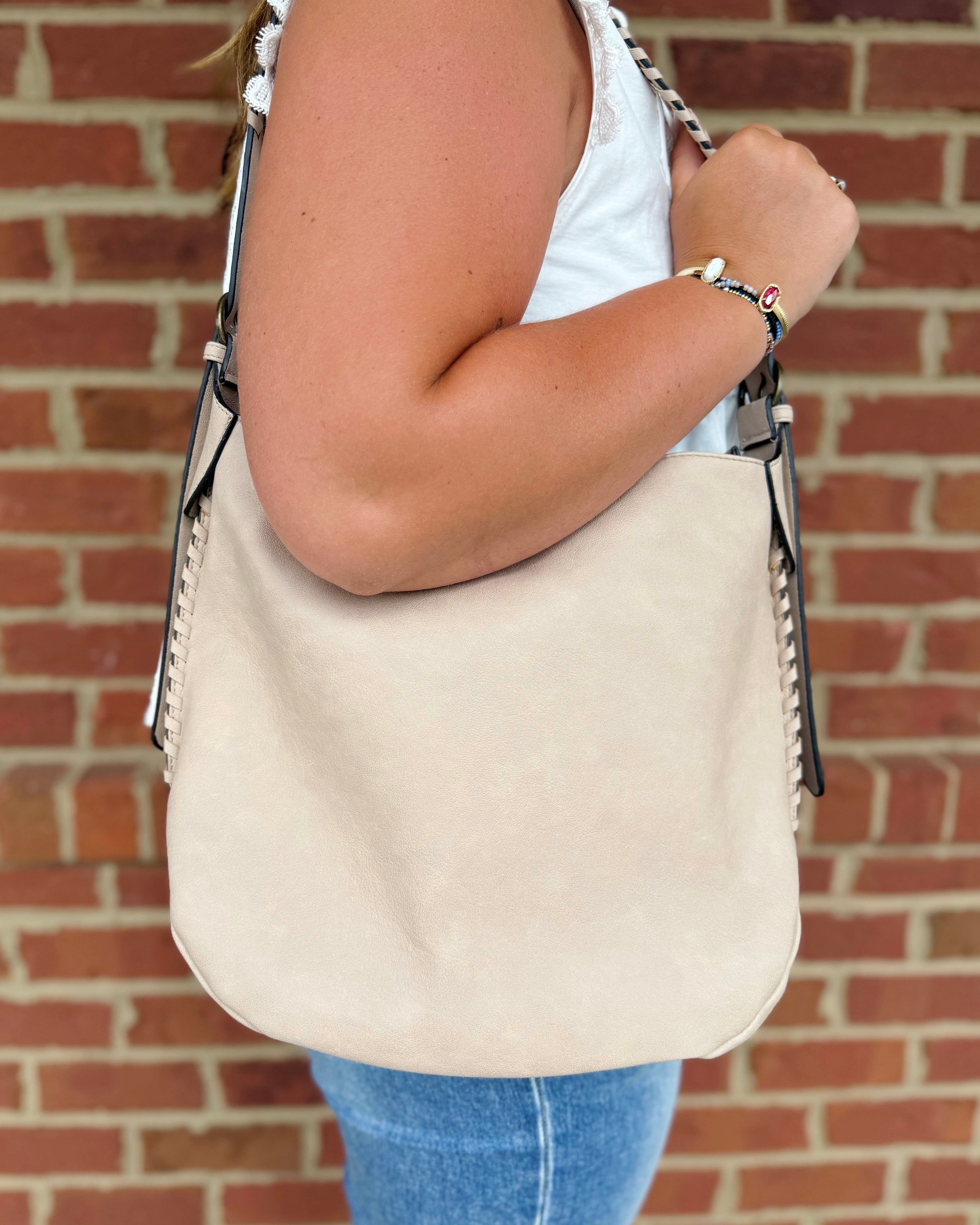 A woman wearing a white tank top and blue jeans carries a large beige suede shoulder bag with black and silver chain strap details, set against a red brick wall background.