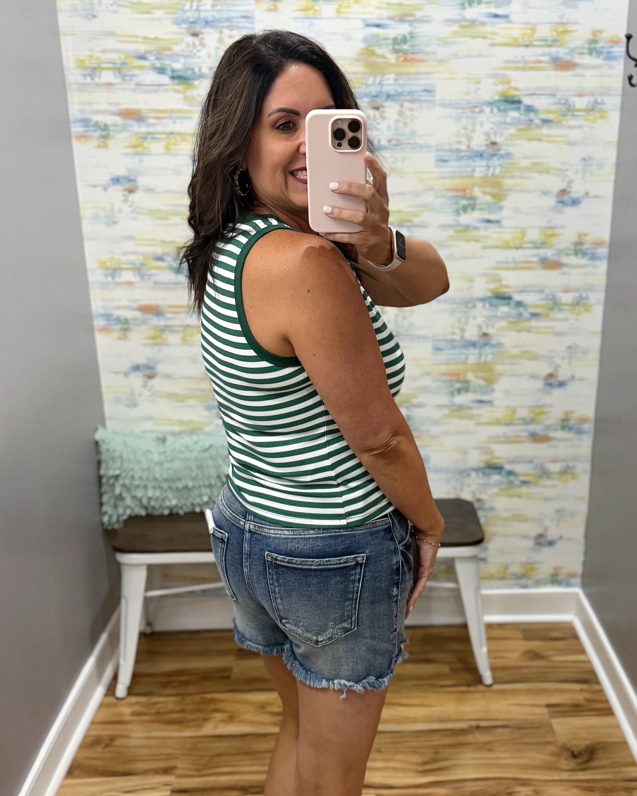 Woman taking a mirror selfie wearing a green and white striped tank top and denim shorts in a room with a textured wall.