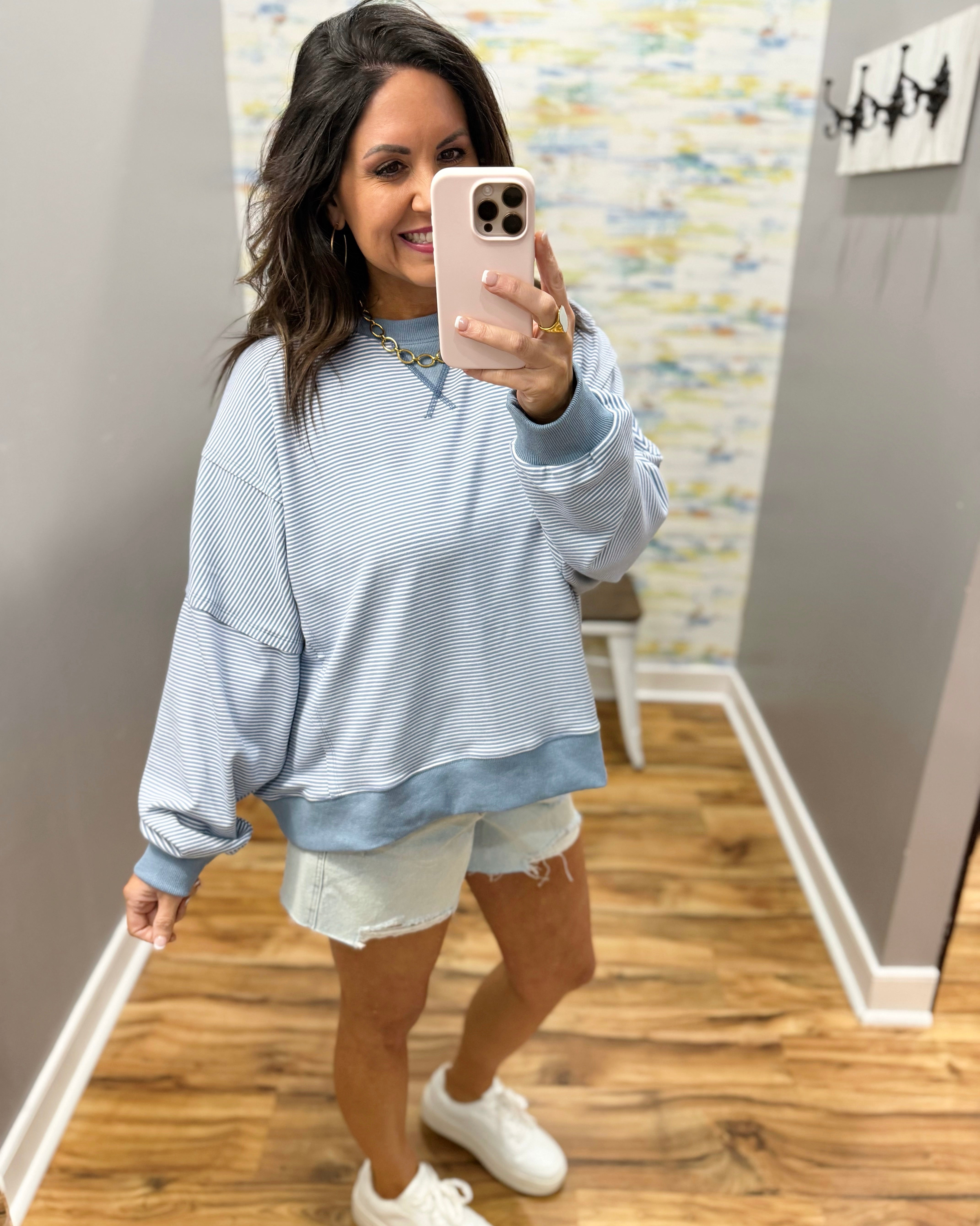 Woman taking a mirror selfie wearing a light blue sweatshirt and white shorts in a room with a textured wall.