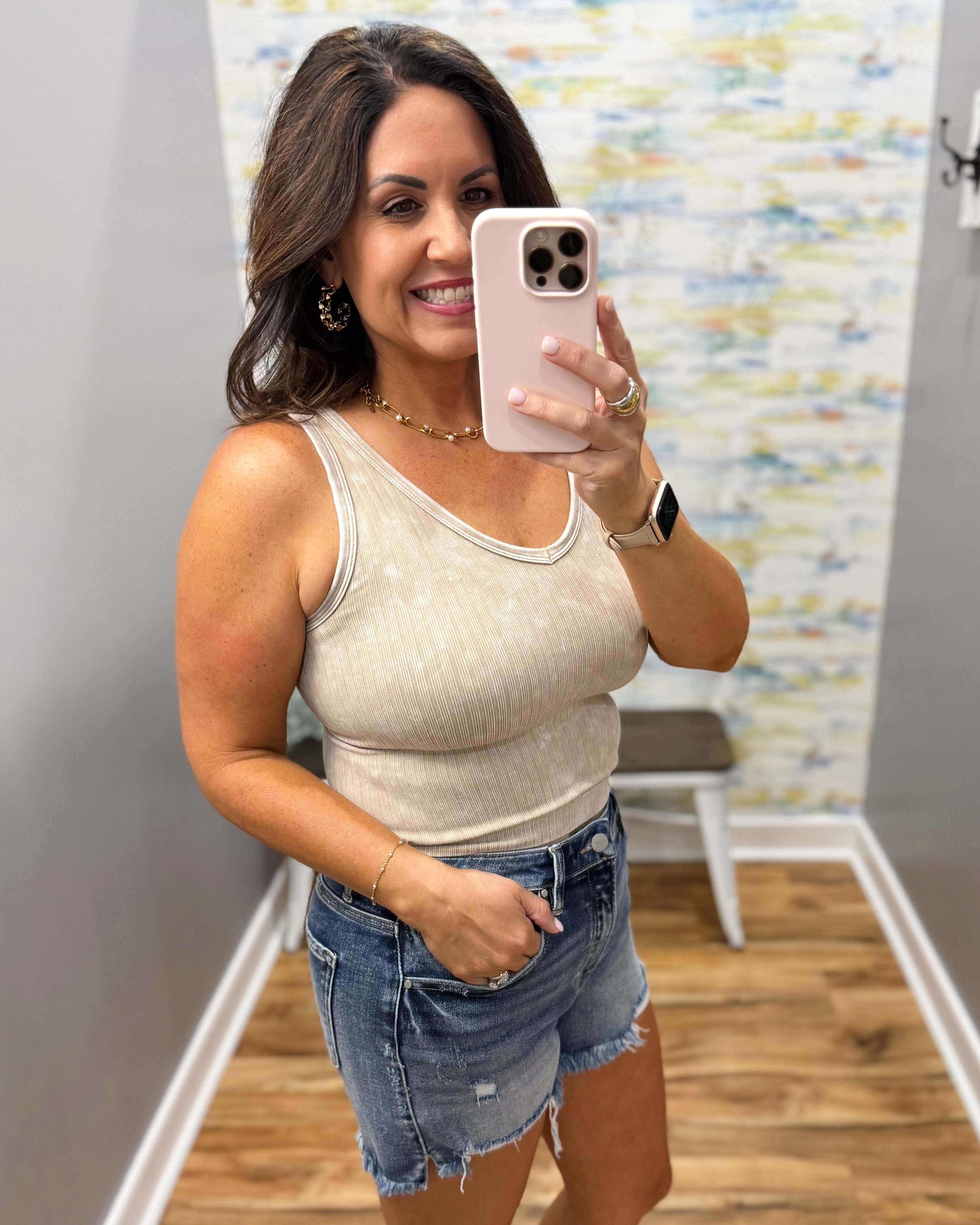 Woman taking a mirror selfie wearing a beige tank top and denim shorts in a room with a colorful wall.