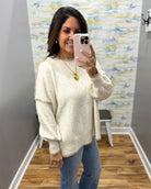 A woman takes a mirror selfie wearing a cream-colored knit sweater with gold trim on the shoulders and a gold pendant necklace, paired with blue jeans.