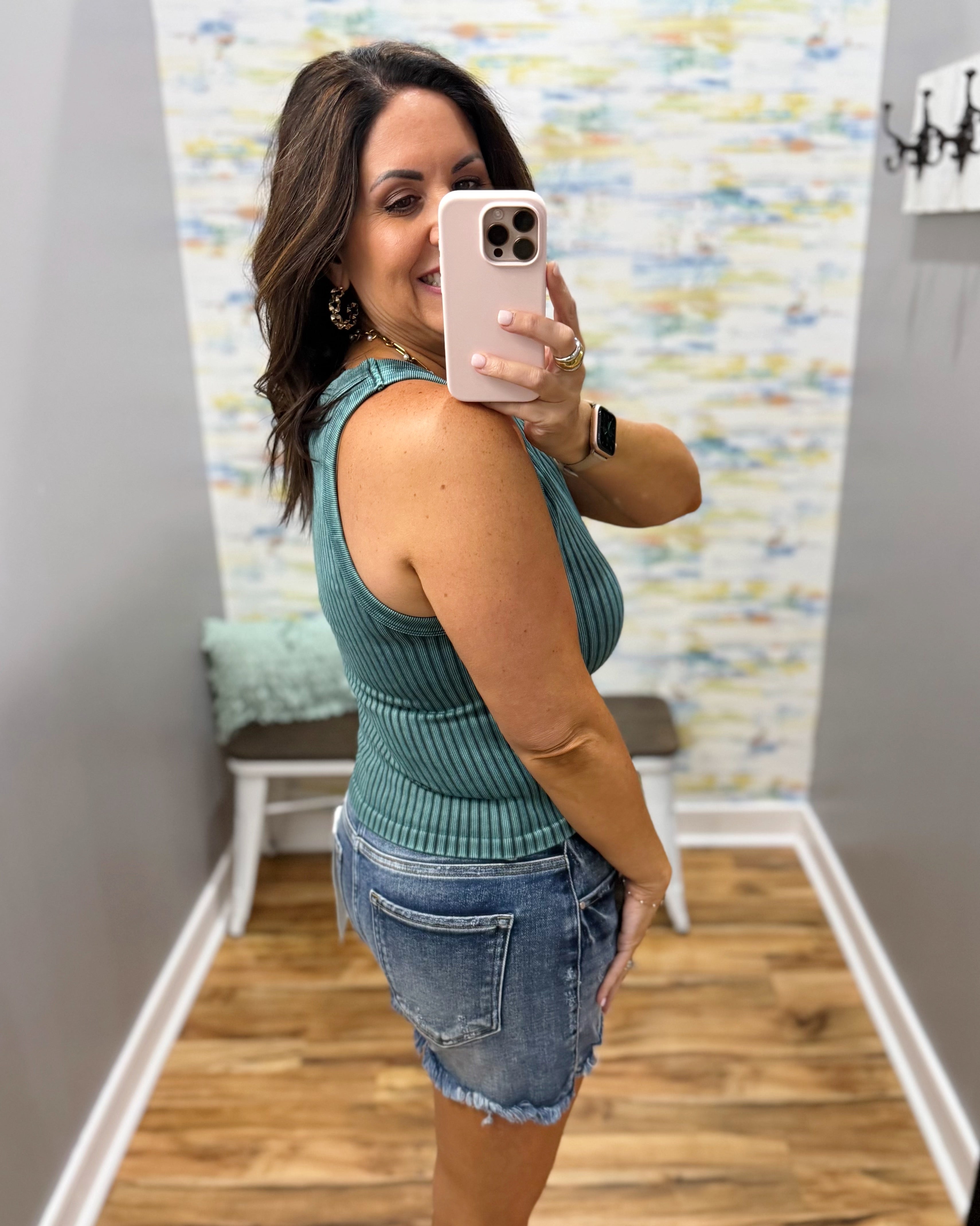 Woman taking a mirror selfie wearing a green ribbed tank top and denim shorts in a room with a patterned wall.