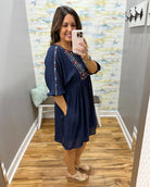 Woman taking a mirror selfie wearing a navy blue dress with colorful embroidery in a room with a patterned wall.