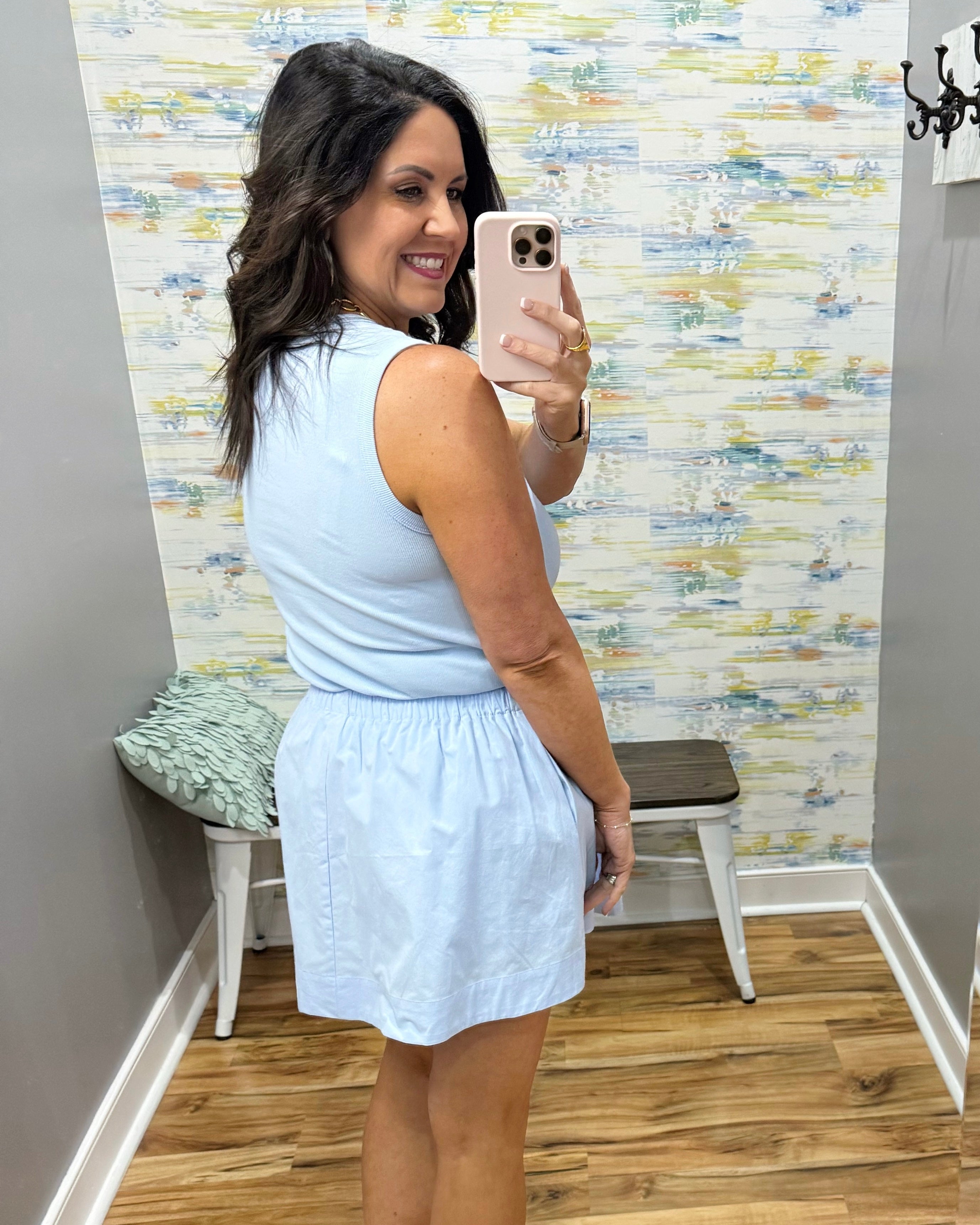 Woman taking a mirror selfie wearing a light blue sleeveless top and white shorts in a room with patterned wallpaper.