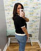 A woman taking a mirror selfie in a fitting room, wearing a black short-sleeve knit top with white contrast trim at the collar, cuffs, and hem, paired with blue jeans.