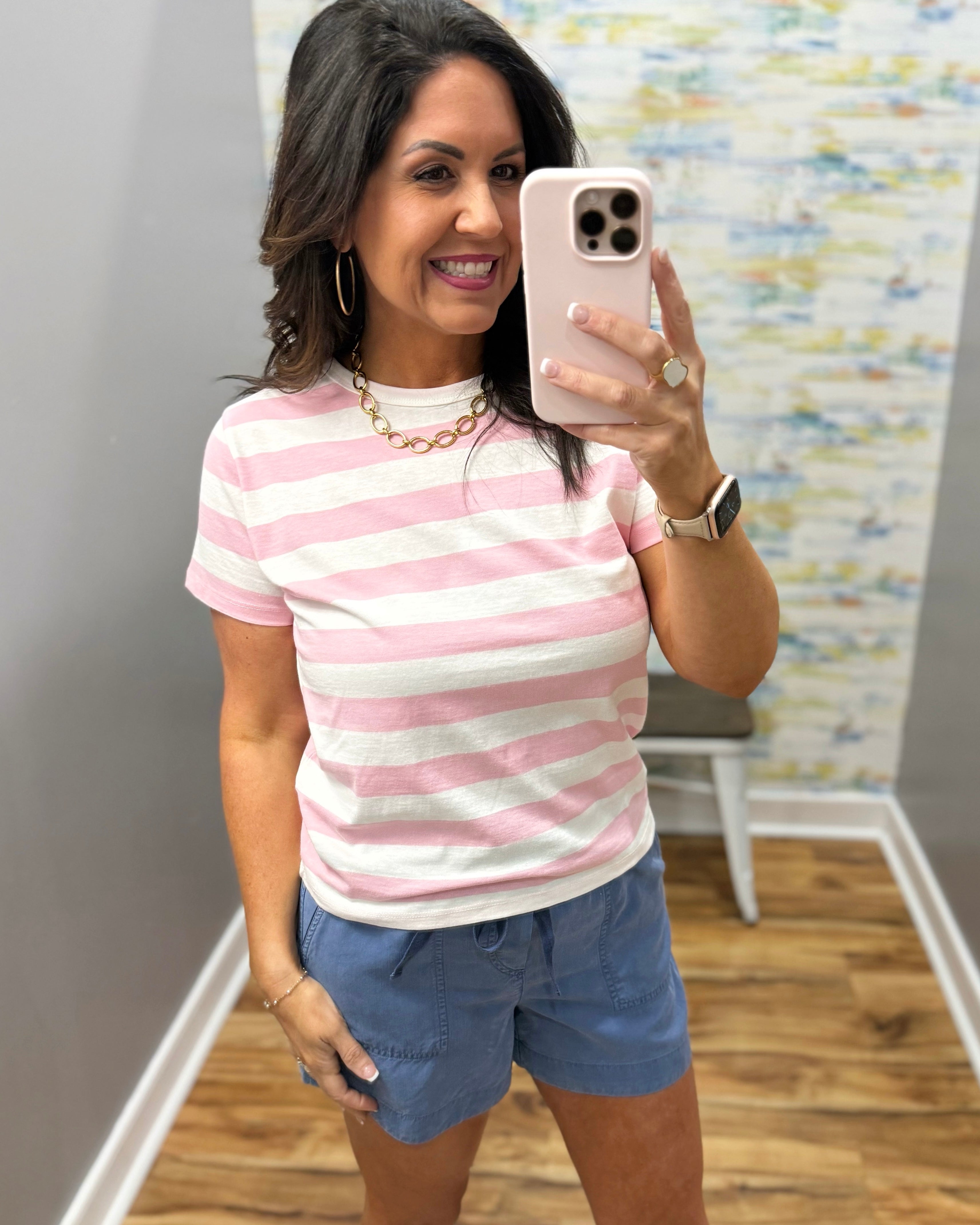 Woman taking a mirror selfie wearing a pink and white striped shirt and blue shorts.
