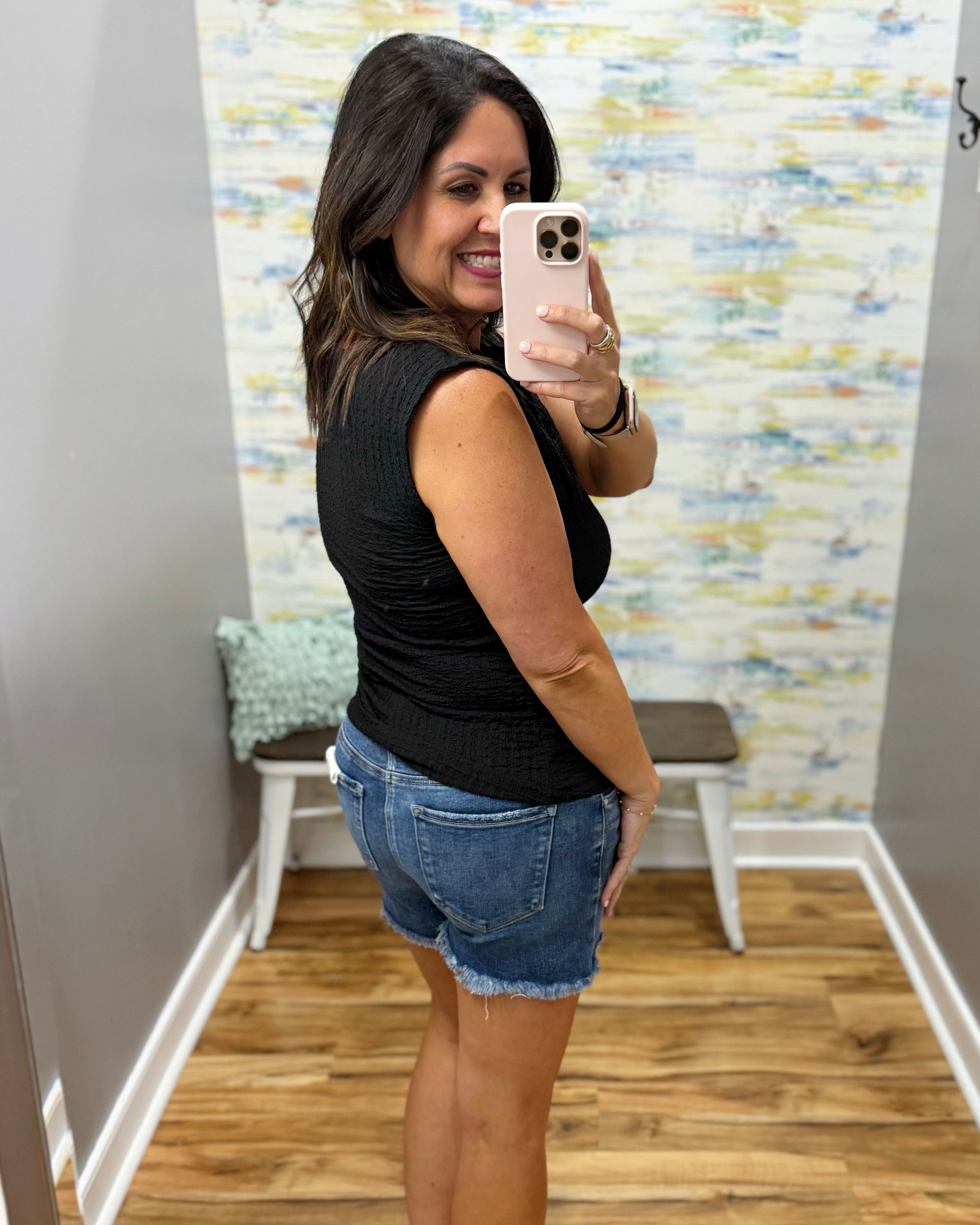 Woman taking a mirror selfie wearing a black sleeveless top and denim shorts in a room with colorful wall art.