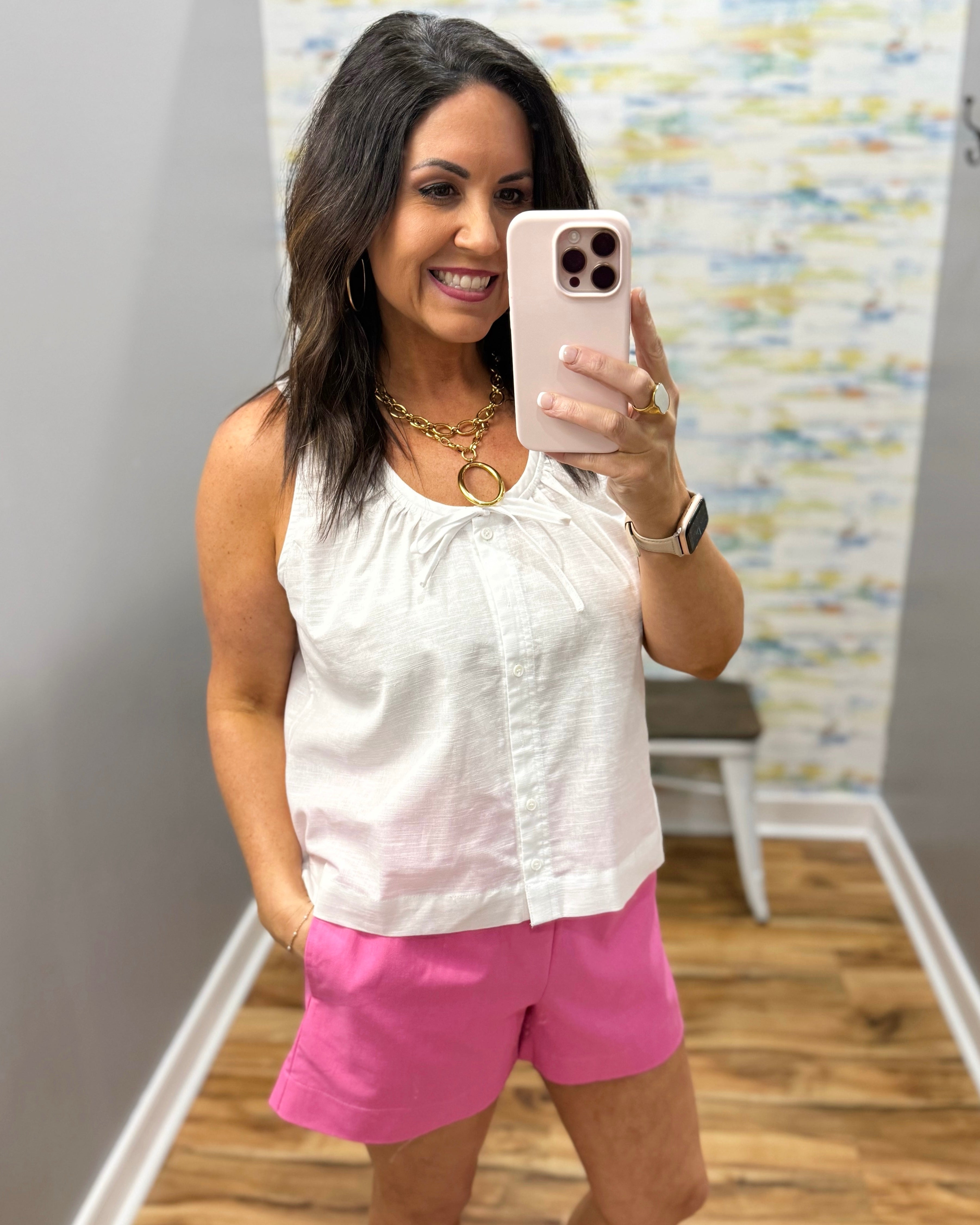 Woman taking a mirror selfie wearing a white sleeveless top and pink shorts.