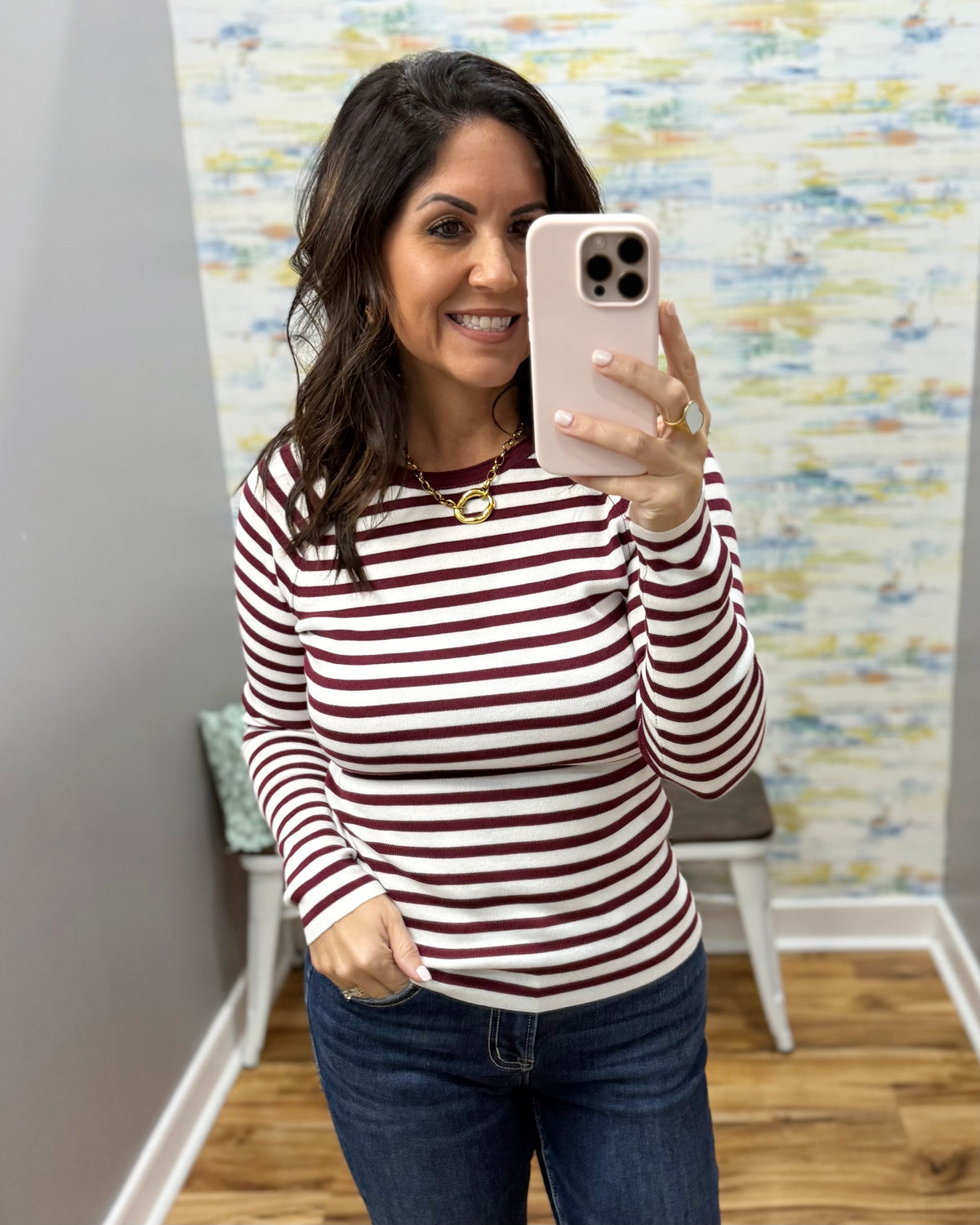 A woman taking a mirror selfie wearing a long-sleeve crewneck top with horizontal burgundy and off-white stripes, paired with dark blue jeans.