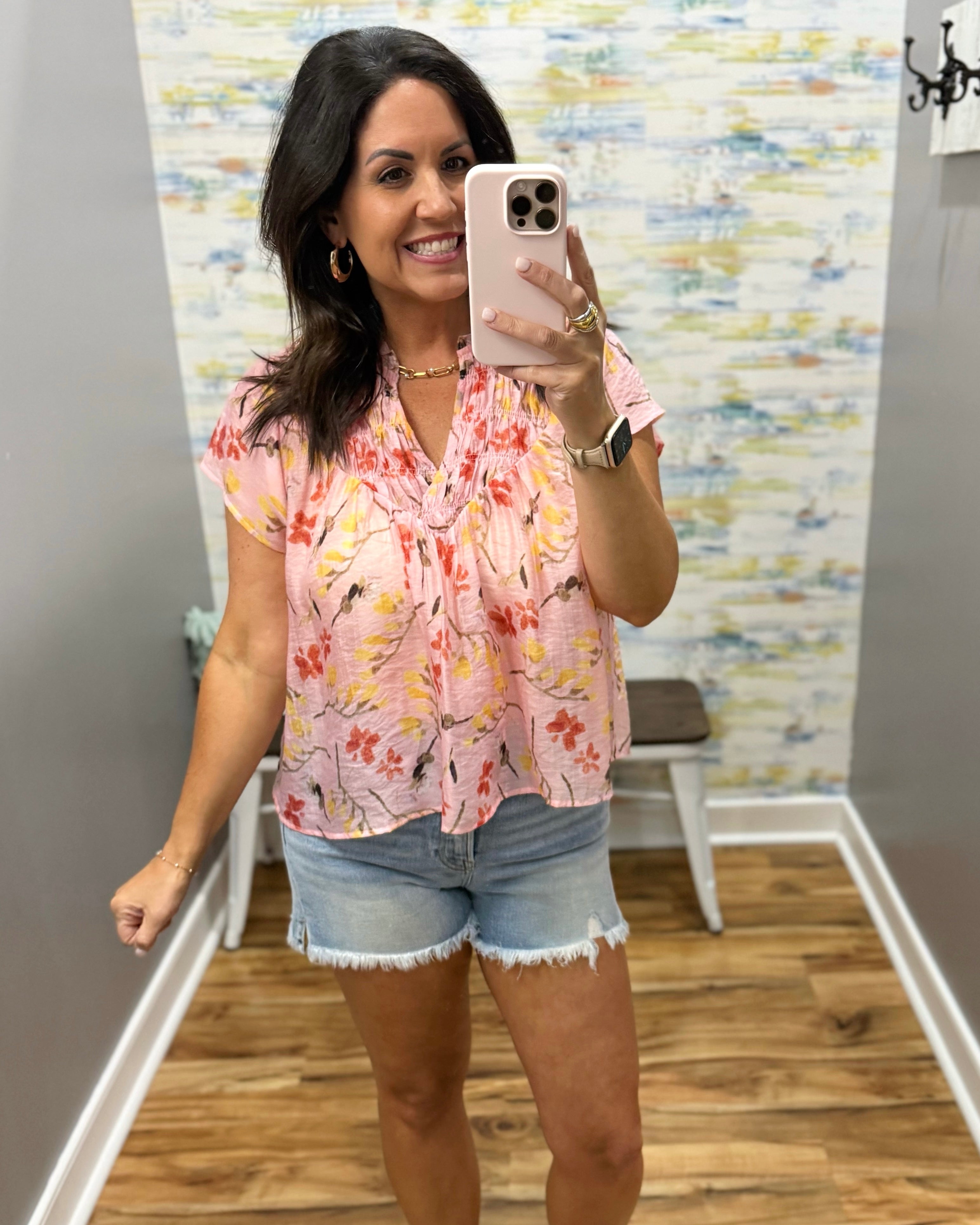 Woman taking a mirror selfie wearing a floral top and denim shorts in a room with a patterned wall.