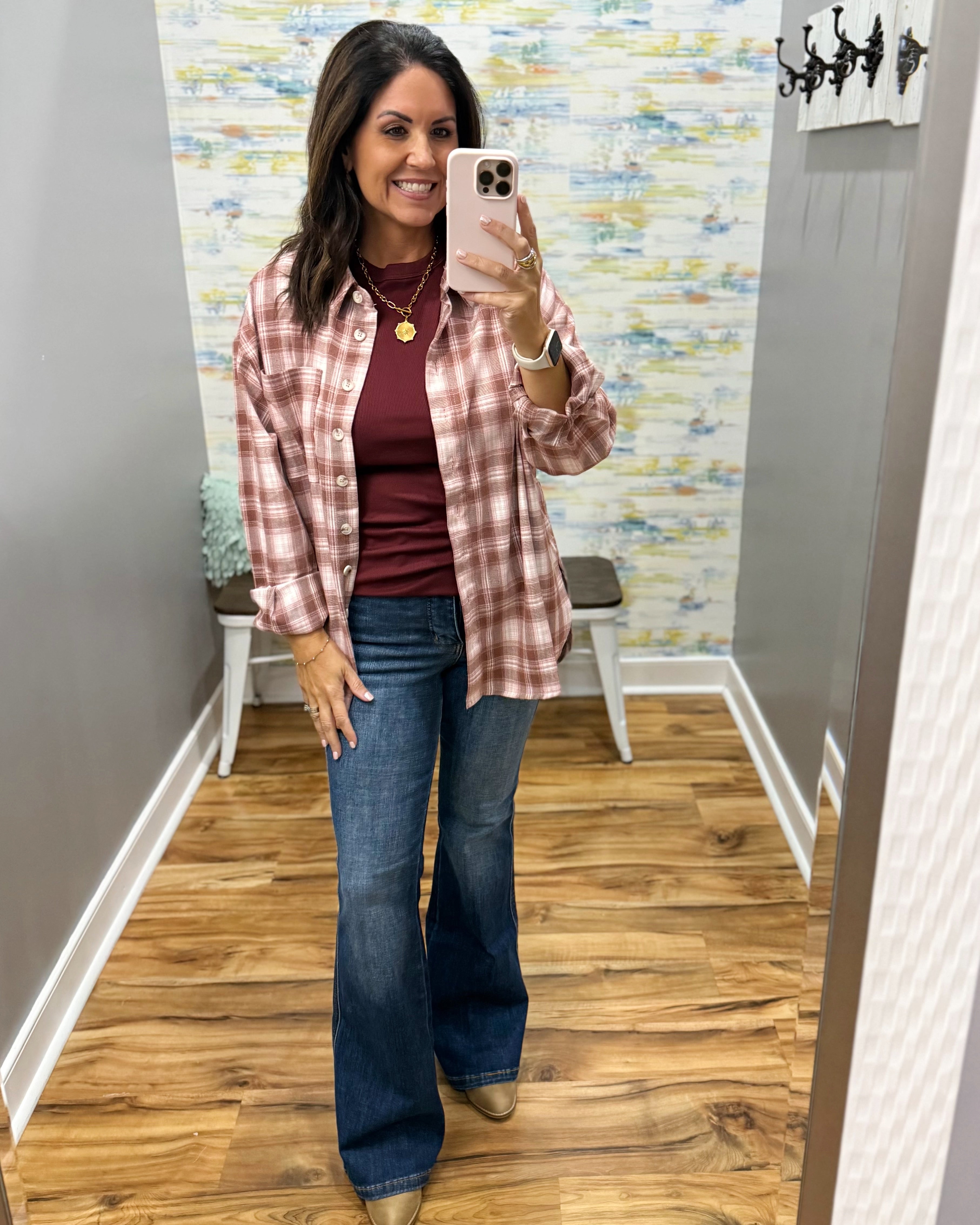 A woman takes a mirror selfie in a fitting room showing a casual outfit consisting of a red and white plaid button-down shirt over a dark red top, blue flare jeans, and tan boots.