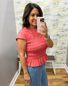 A woman takes a mirror selfie wearing a coral pink short-sleeve smocked peplum top and blue jeans in a fitting room.