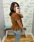 A woman takes a mirror selfie in a boutique fitting room wearing a rust-brown short-sleeve smocked top with a ruffled hem and light-wash blue jeans.