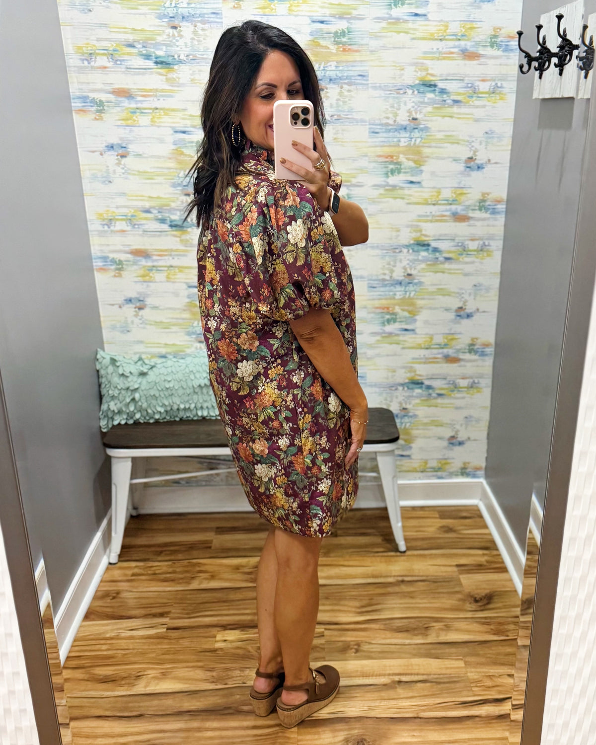 A woman takes a mirror selfie in a fitting room wearing a short-sleeve floral dress with a high collar. The dress has a burgundy base with yellow, orange, and white floral patterns.