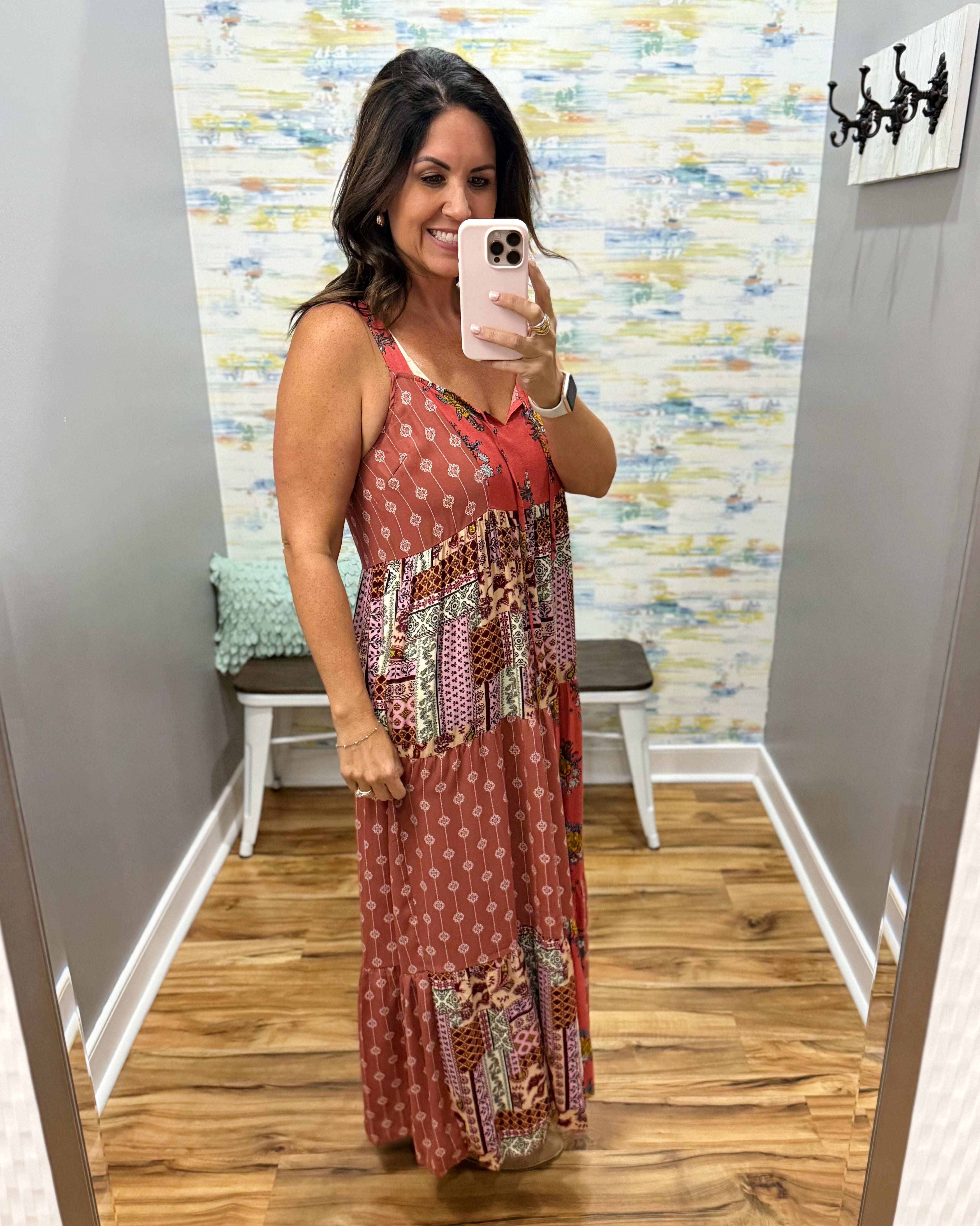 A woman takes a mirror selfie in a fitting room wearing a sleeveless patchwork maxi dress with a rust red base and multicolor geometric patterns. The background features gray walls, a patterned wallpaper, a wooden floor, and a small bench.