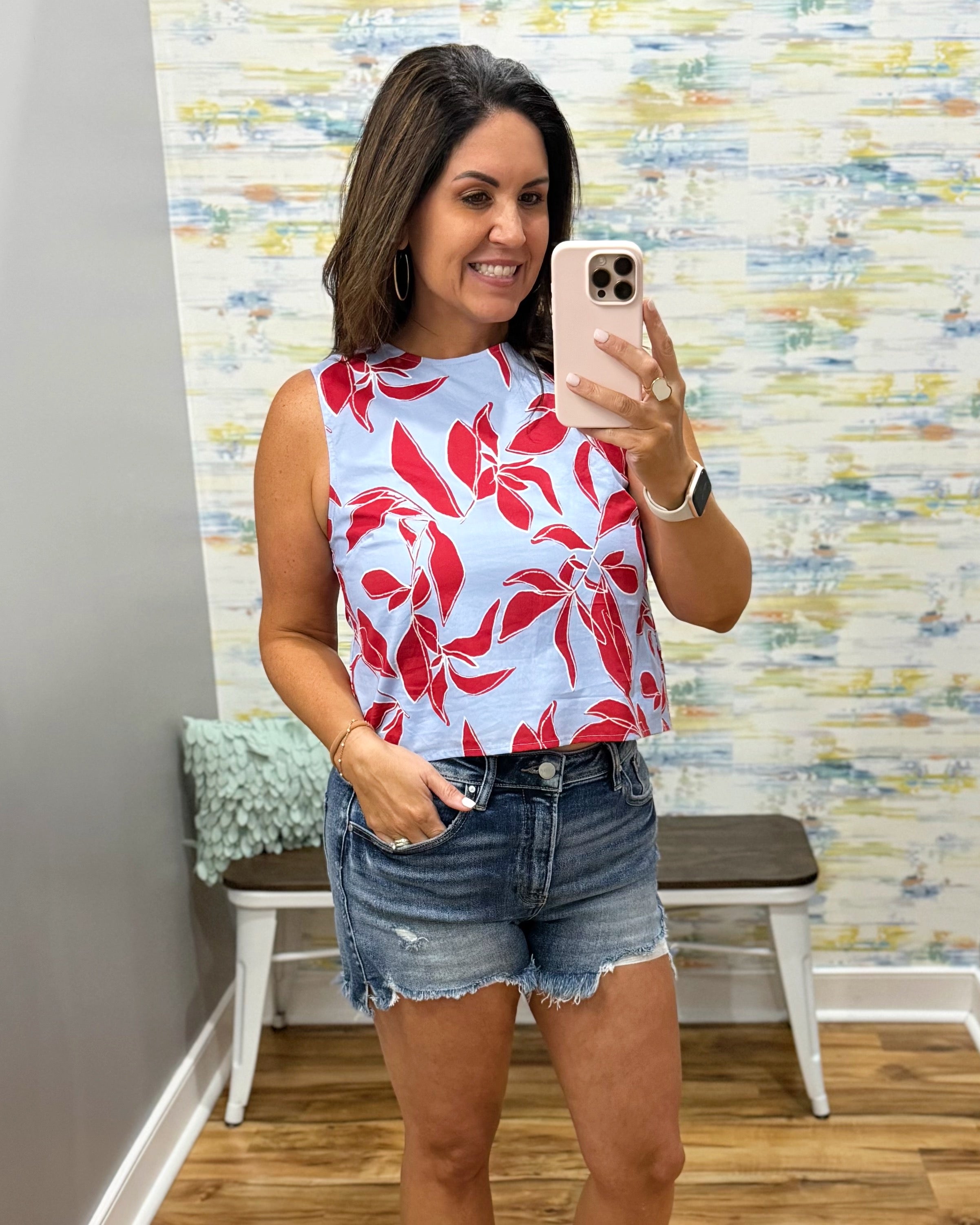 Woman taking a mirror selfie wearing a red floral top and denim shorts in a room with a colorful wall.