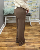 A person wearing high-waisted, wide-leg brown pants and a cream long-sleeve top, standing on a wood floor in front of a bench and patterned wall.