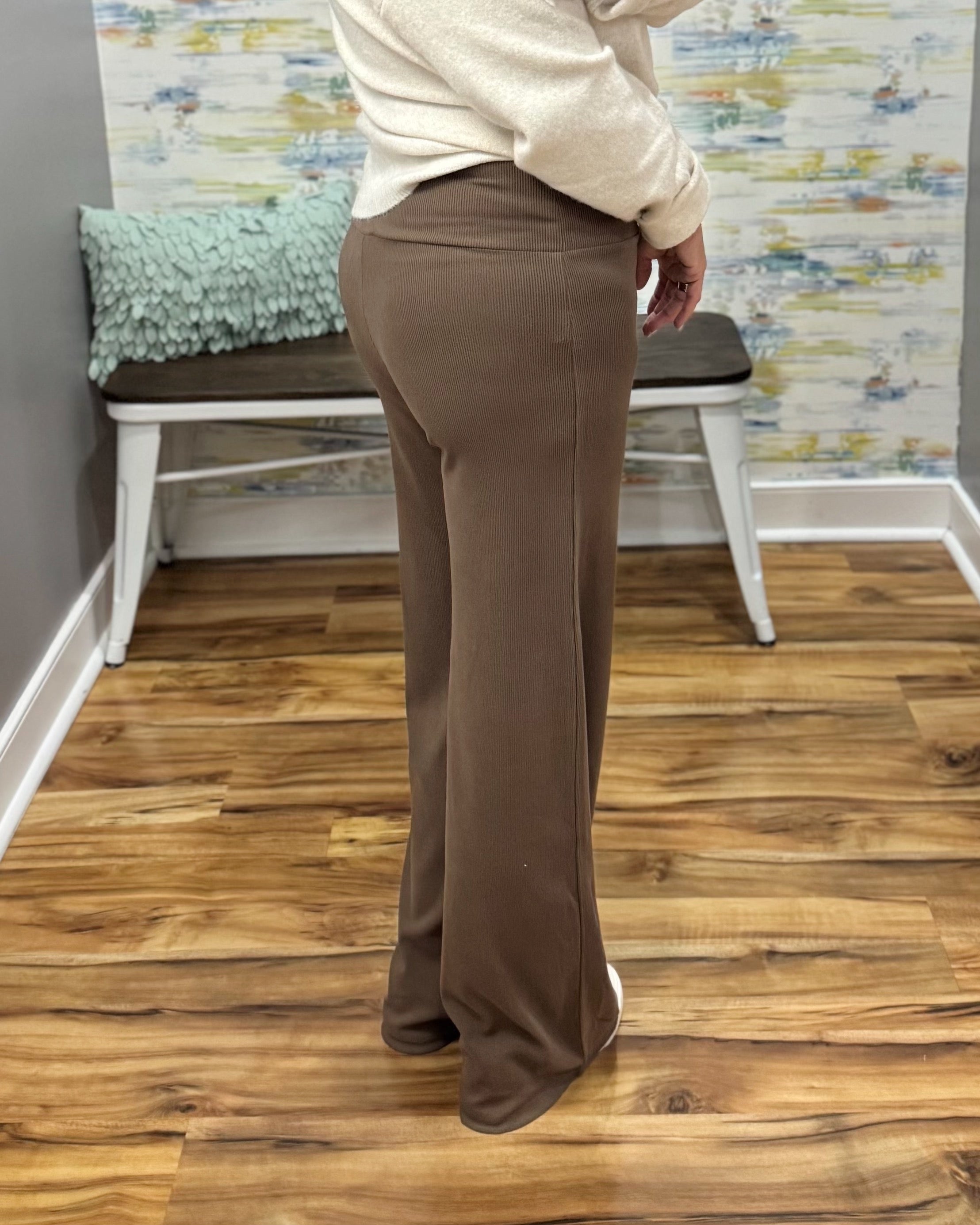 A person wearing high-waisted, wide-leg brown pants and a cream long-sleeve top, standing on a wood floor in front of a bench and patterned wall.