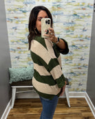 A woman takes a mirror selfie wearing a ribbed knit sweater with wide green and cream stripes, paired with blue jeans, in front of a patterned wallpapered wall.