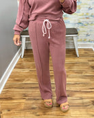 A person wearing a dusty rose pink sweatshirt and matching sweatpants with a white drawstring, standing on a wood floor in an indoor room.