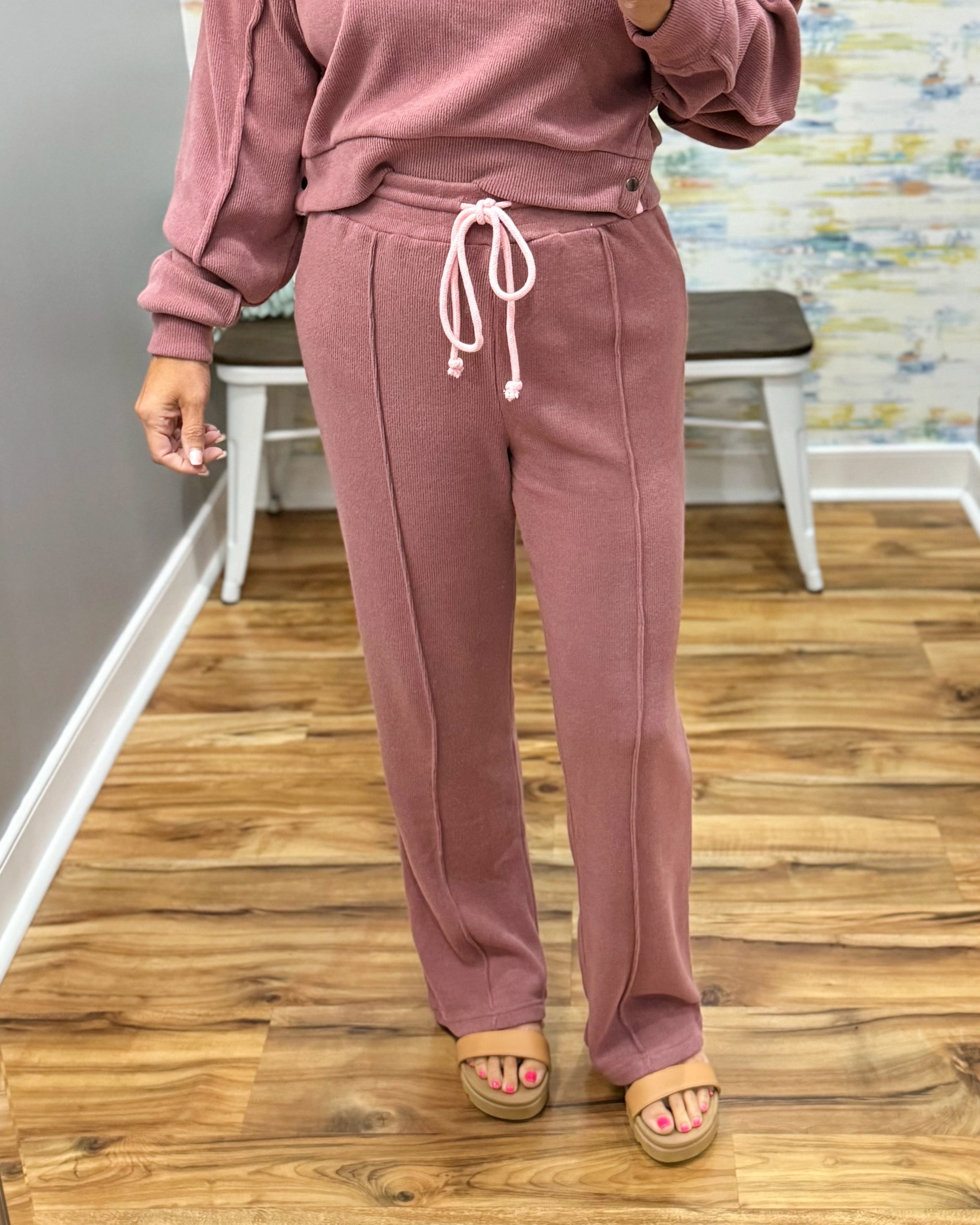 A person wearing a dusty rose pink sweatshirt and matching sweatpants with a white drawstring, standing on a wood floor in an indoor room.