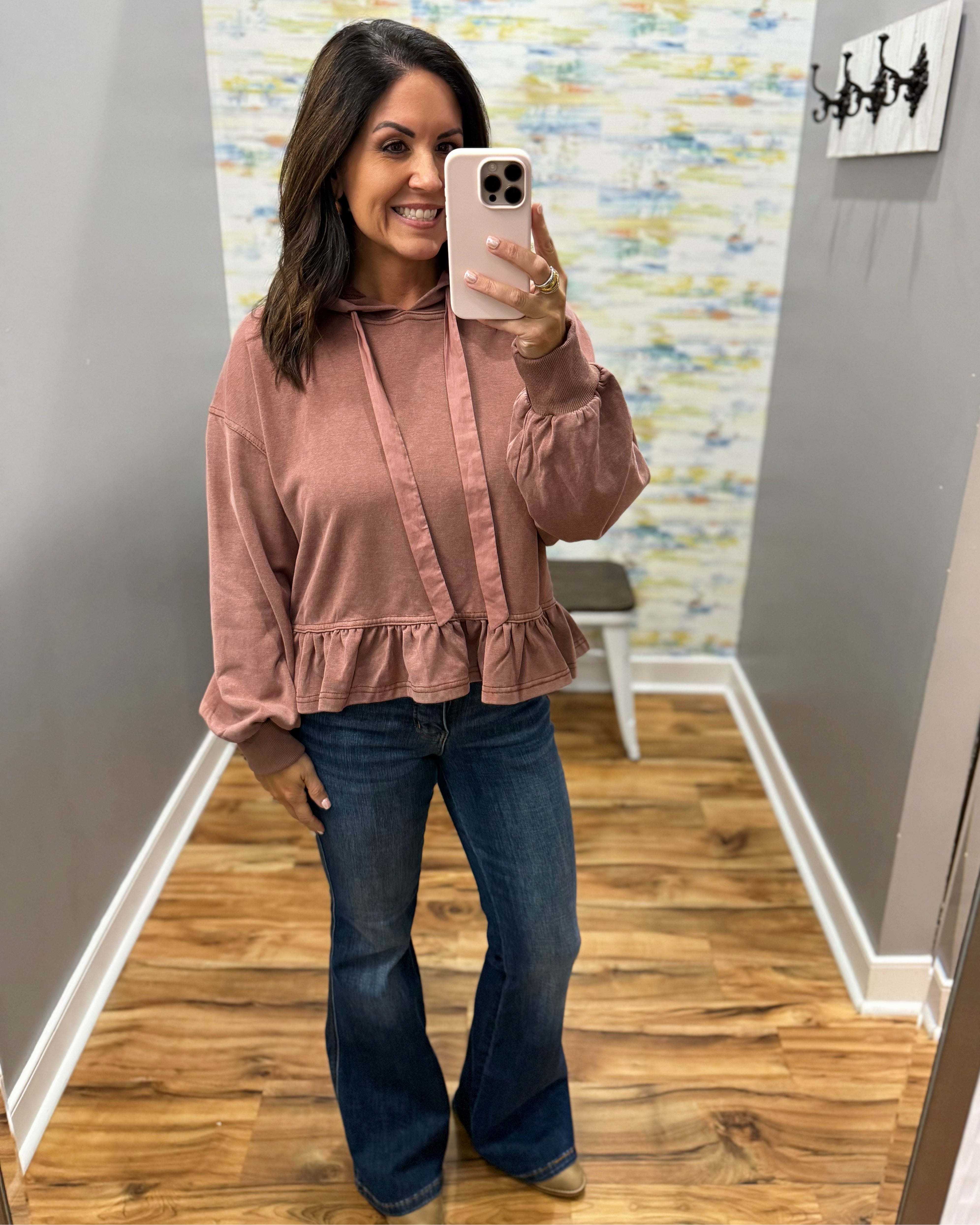 A woman takes a mirror selfie wearing a dusty rose long-sleeve ruffled blouse and dark blue flare jeans in a home hallway.
