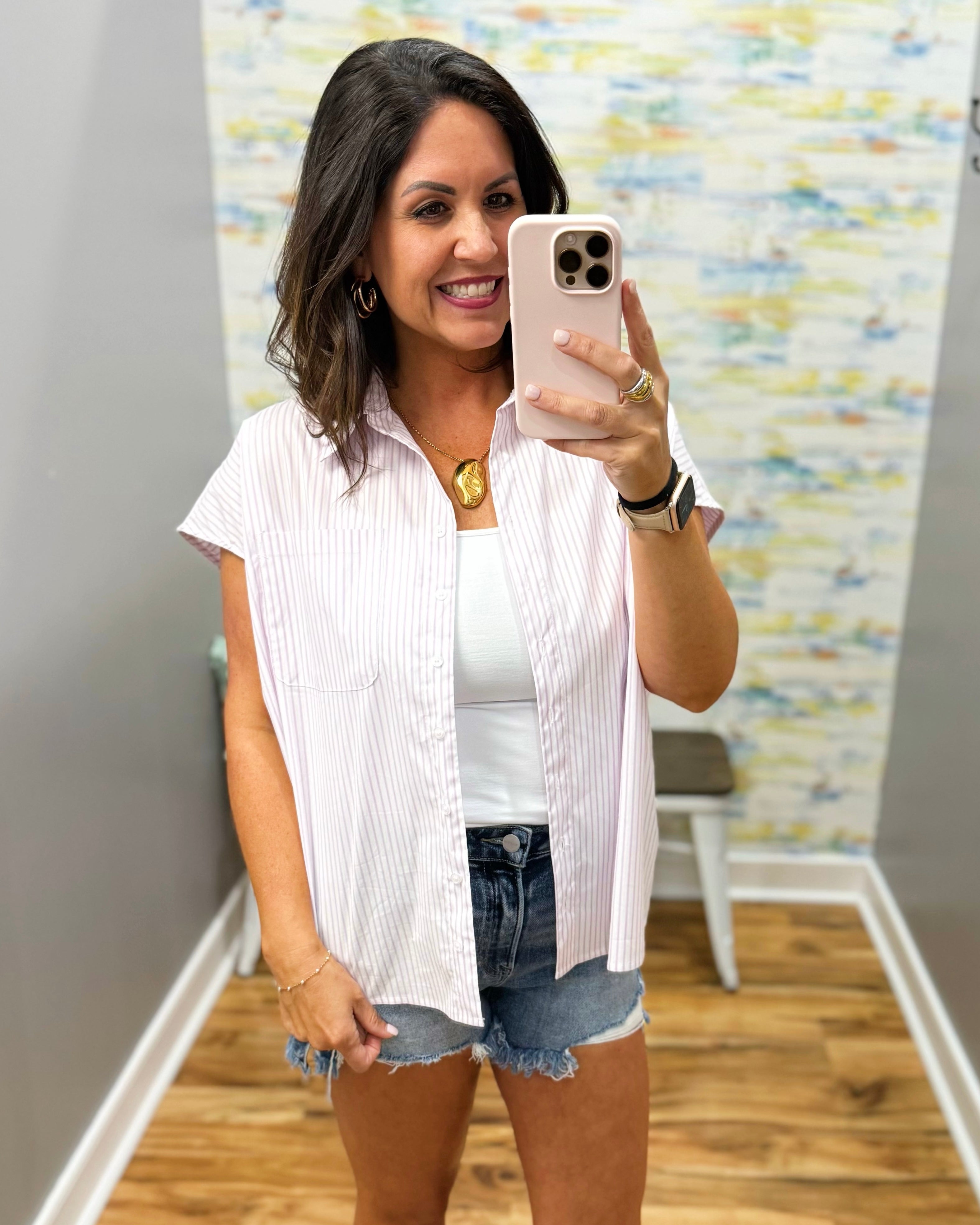 Woman taking a mirror selfie wearing a white button-up shirt and denim shorts in a room with a colorful wall.