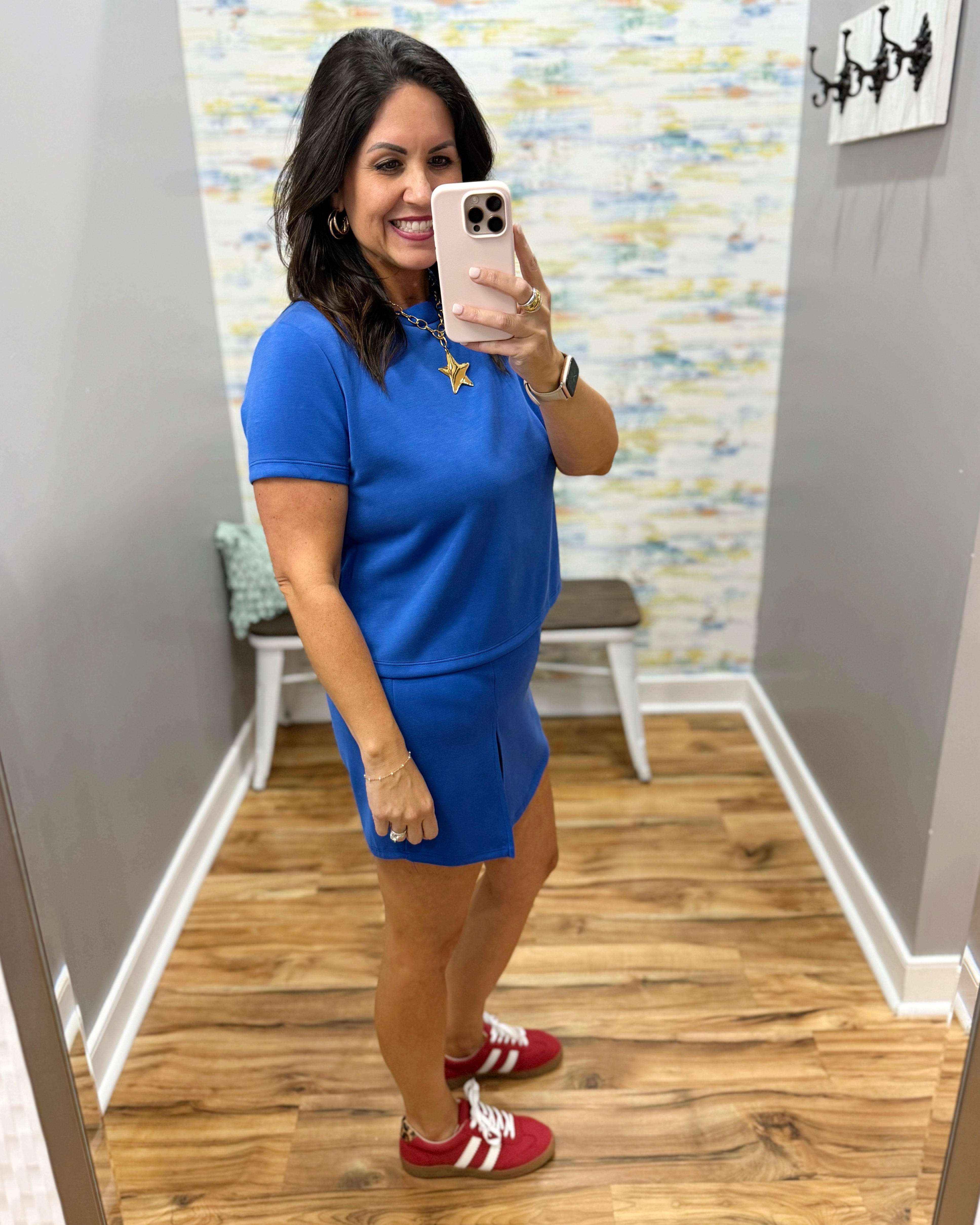 Woman taking a mirror selfie wearing a blue dress and red sneakers in a room with a patterned wall.