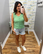 Woman taking a mirror selfie wearing a green sleeveless top and denim shorts in a room with patterned wallpaper.