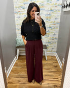 A woman takes a mirror selfie in a fitting room wearing a black short-sleeve sweater and high-waisted wide-leg pants in merlot with Corkys Sober Up shoes in leopard.
