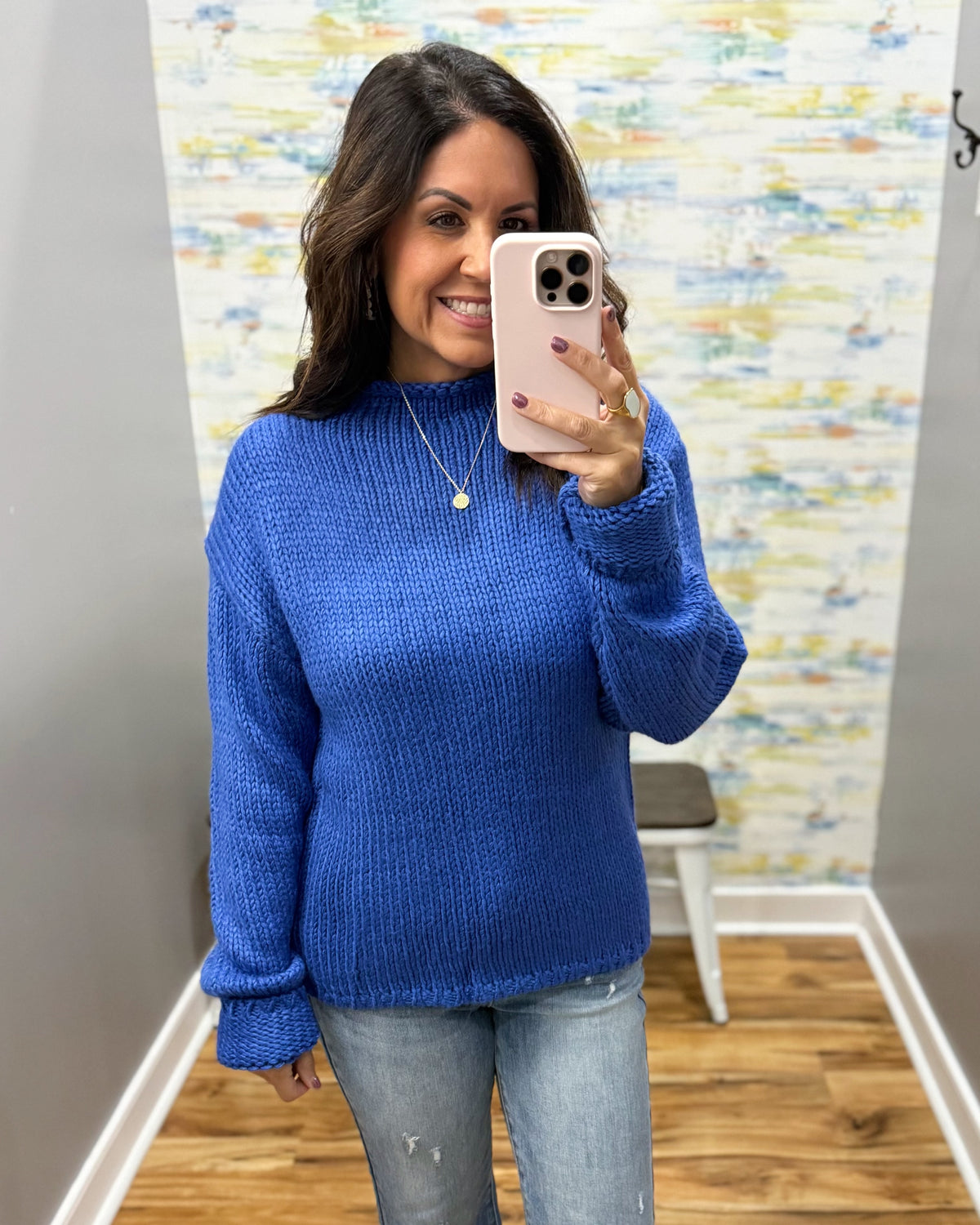 A woman takes a mirror selfie wearing a vibrant royal blue chunky knit sweater and light-wash jeans in a fitting room with a patterned wall and wood floor.