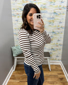 A woman takes a mirror selfie wearing a brown and white horizontally striped long-sleeve top and dark blue jeans in a fitting room with a patterned wall and wood floor.