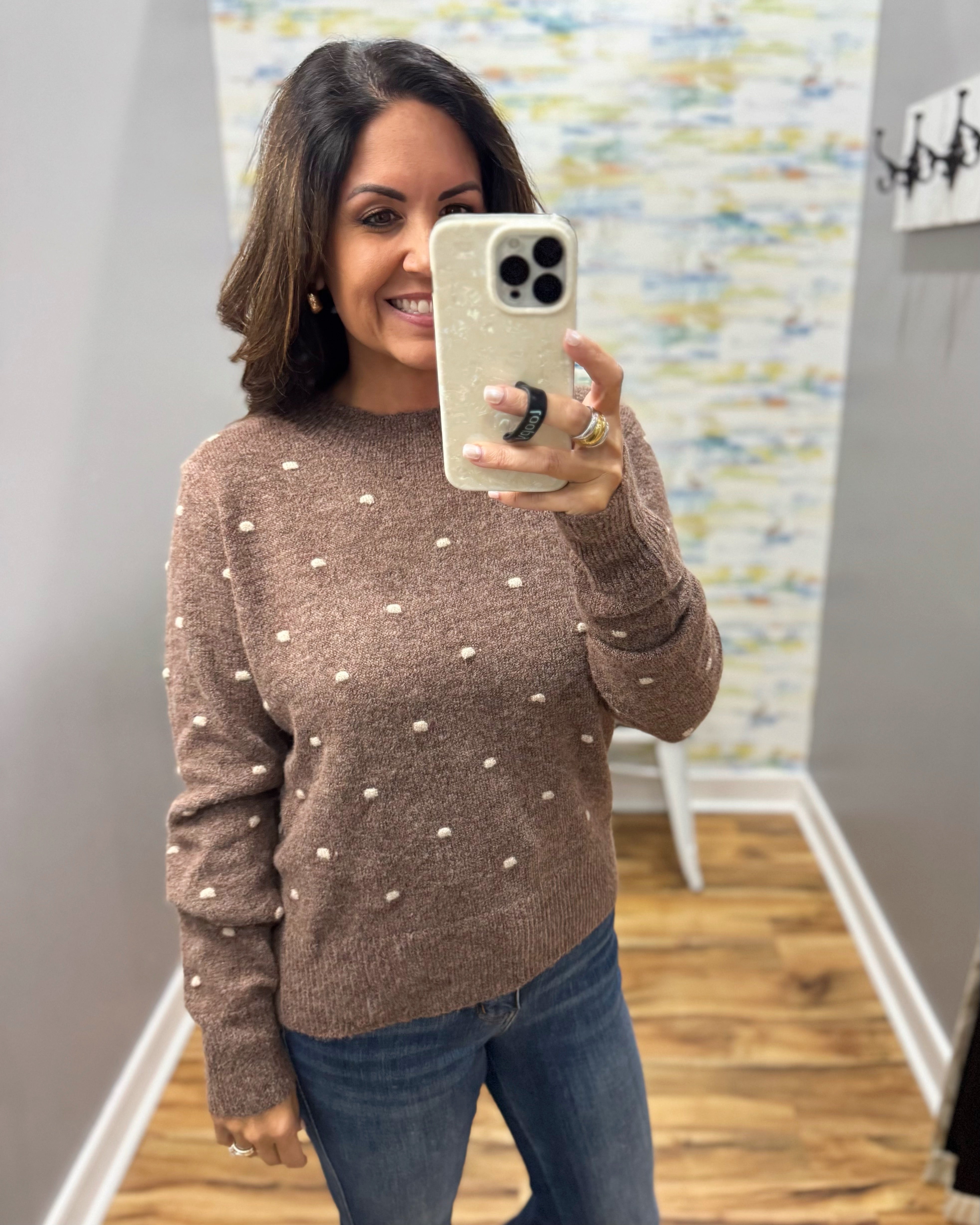 A woman takes a mirror selfie wearing a brown knit sweater with small white pom-pom details and blue jeans in an indoor hallway.