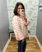 A woman takes a mirror selfie wearing a pink and cream striped knit sweater, blue jeans, and gold jewelry in a home hallway.